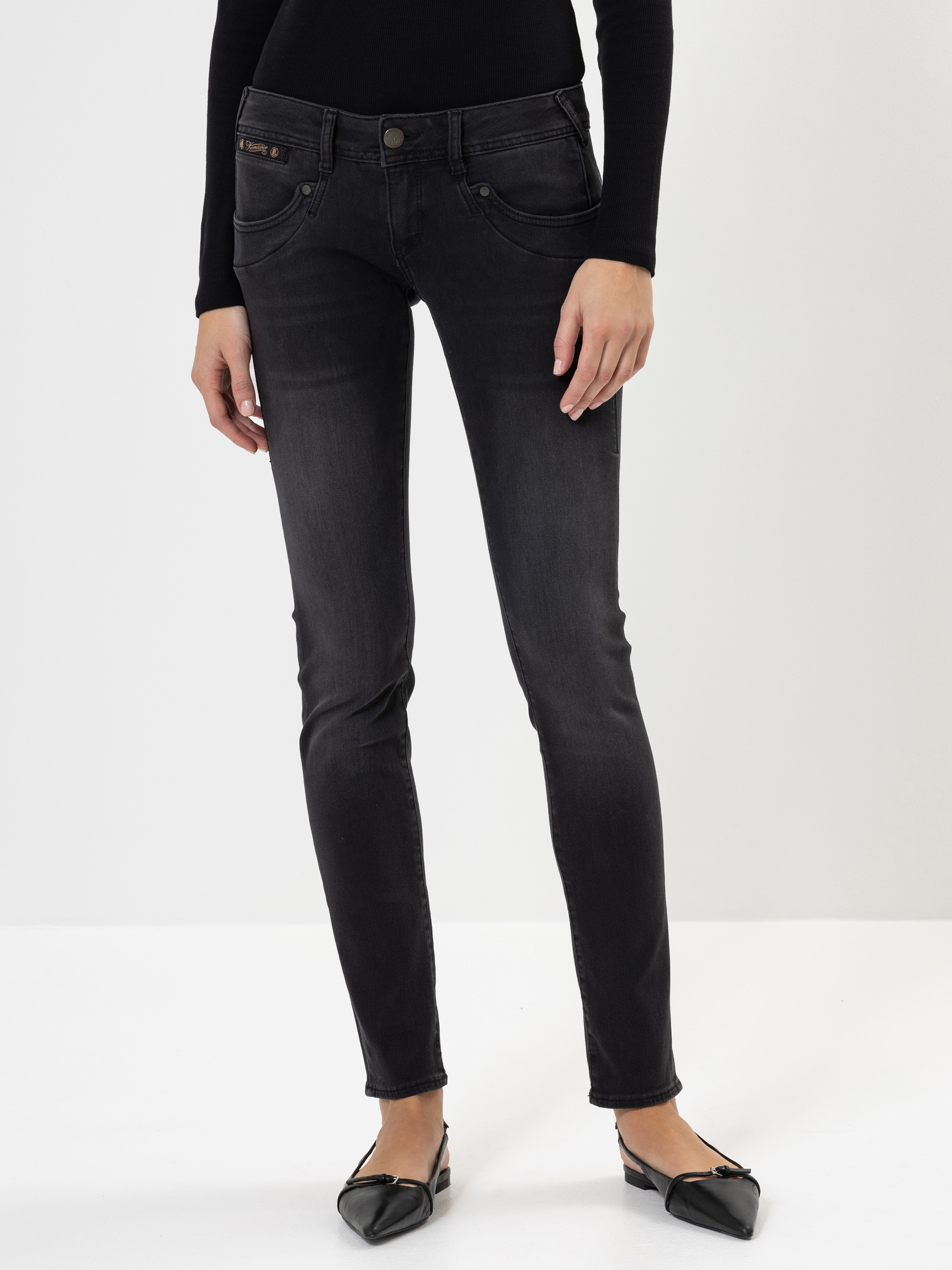 Eine Person, die ein schwarzes, langärmeliges Oberteil, verblasste schwarze Skinny Jeans und schwarze, spitze Schuhe trägt, steht vor einem schlichten weißen Hintergrund.