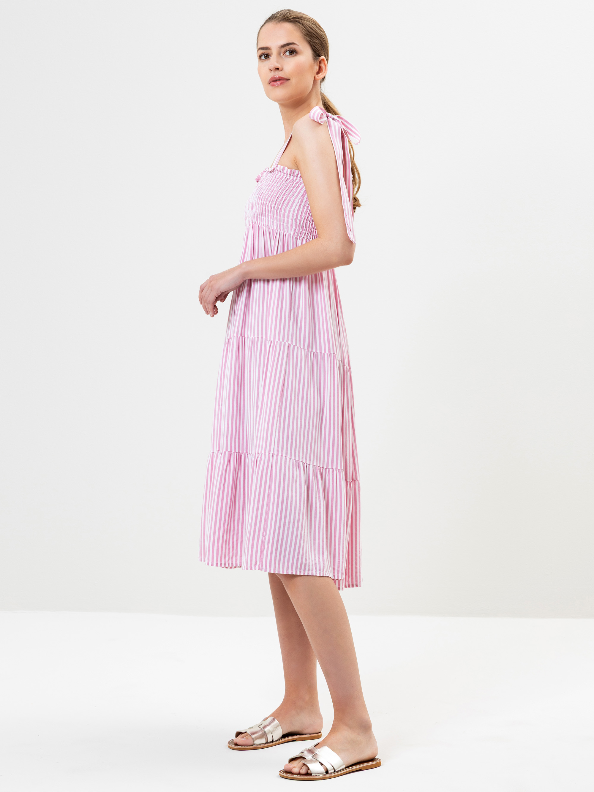 Eine Frau steht seitlich und trägt ein rosa und weiß gestreiftes, ärmelloses Midikleid mit gesmoktem Oberteil und Bindebändern. Sie hat ihr hellbraunes Haar zurückgebunden und trägt weiße flache Sandalen. Der Hintergrund ist einfarbig weiß.