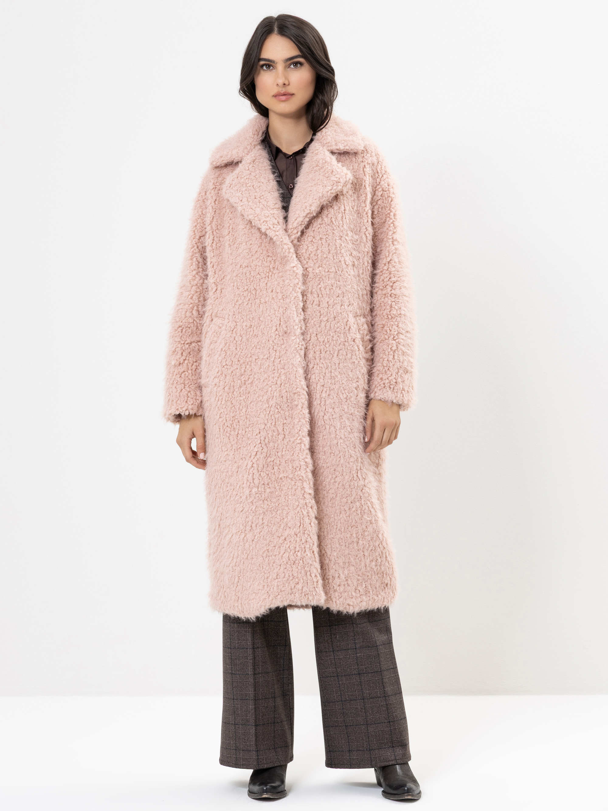 Eine Frau steht vor einem weißen Hintergrund und trägt einen langen, hellrosa, wuscheligen Mantel über einer braunen Bluse und eine braun karierte Hose, dazu schwarze Schuhe. Sie hat schulterlanges dunkles Haar und einen neutralen Gesichtsausdruck.