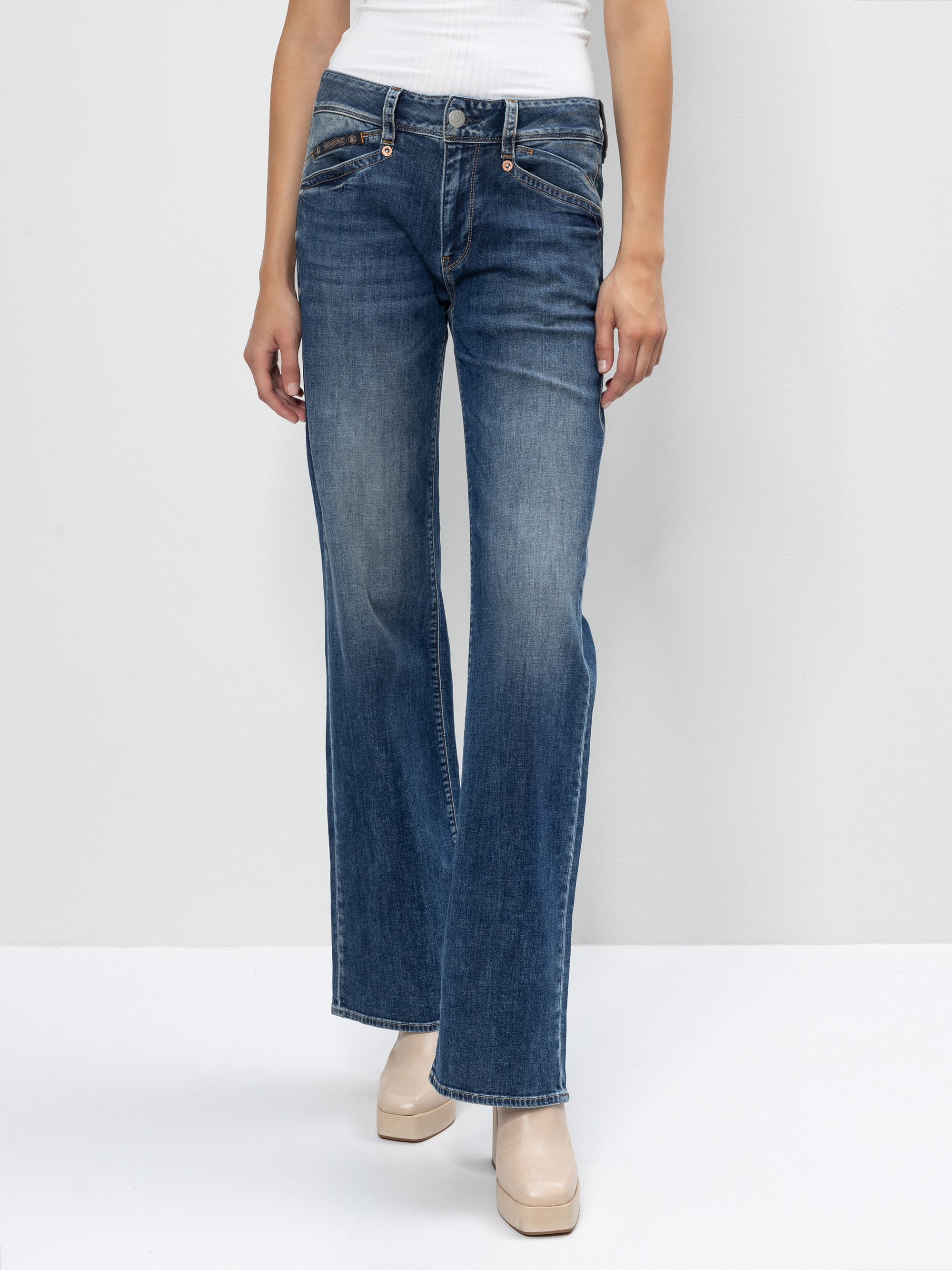 Eine Person, die blaue Schlagjeans und ein weißes geripptes Oberteil trägt, steht vor einem einfarbigen hellen Hintergrund. Nur der untere Teil des Oberkörpers und die Beine sind sichtbar. Die Person trägt außerdem beigefarbene Plateauschuhe.