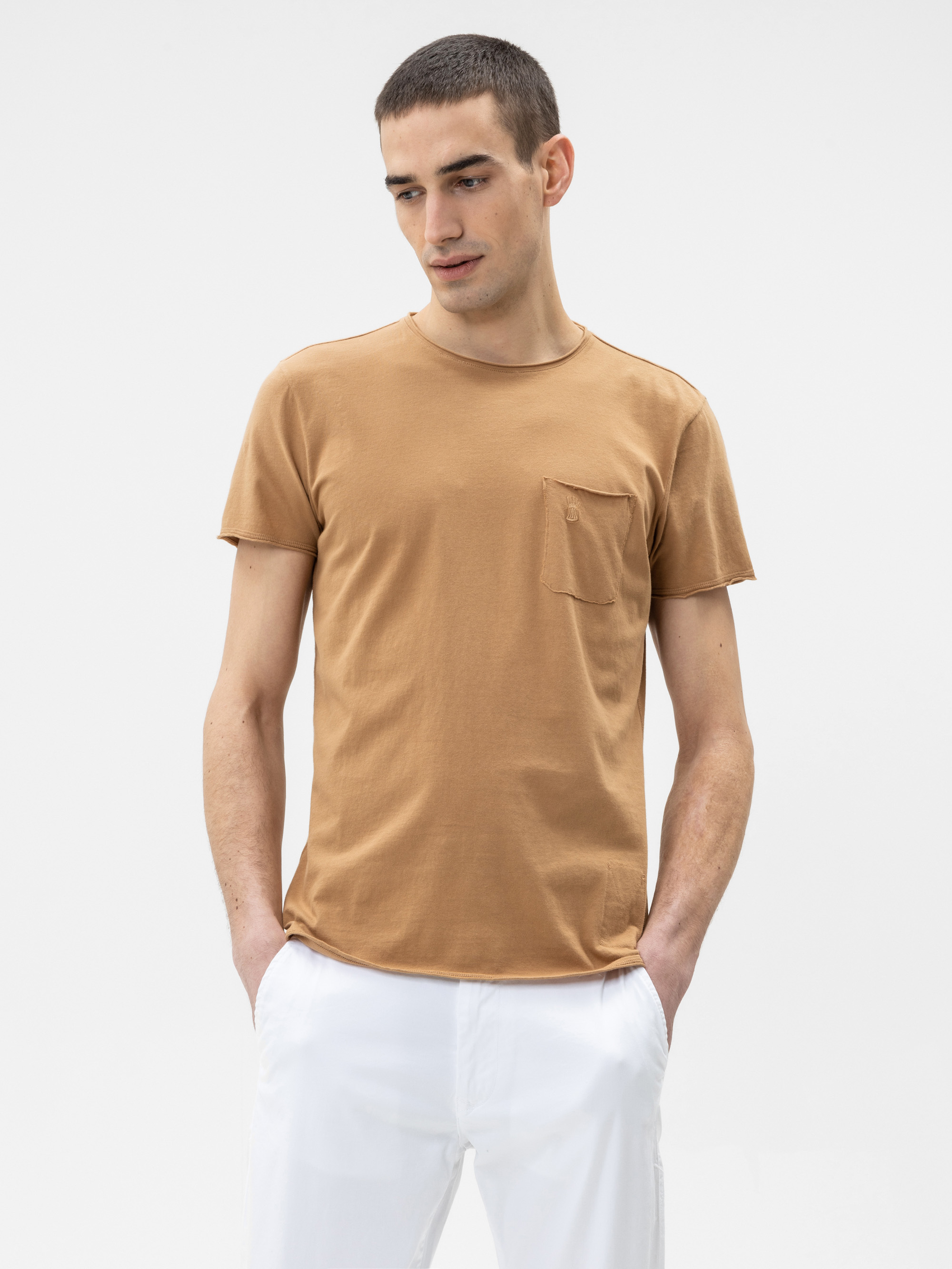 Ein Mann mit kurzen braunen Haaren, der ein hellbraunes T-Shirt mit einer kleinen Brusttasche und eine weiße Hose trägt, steht vor einem schlichten weißen Hintergrund und schaut leicht nach unten und zur Seite, die Hände in den Taschen.