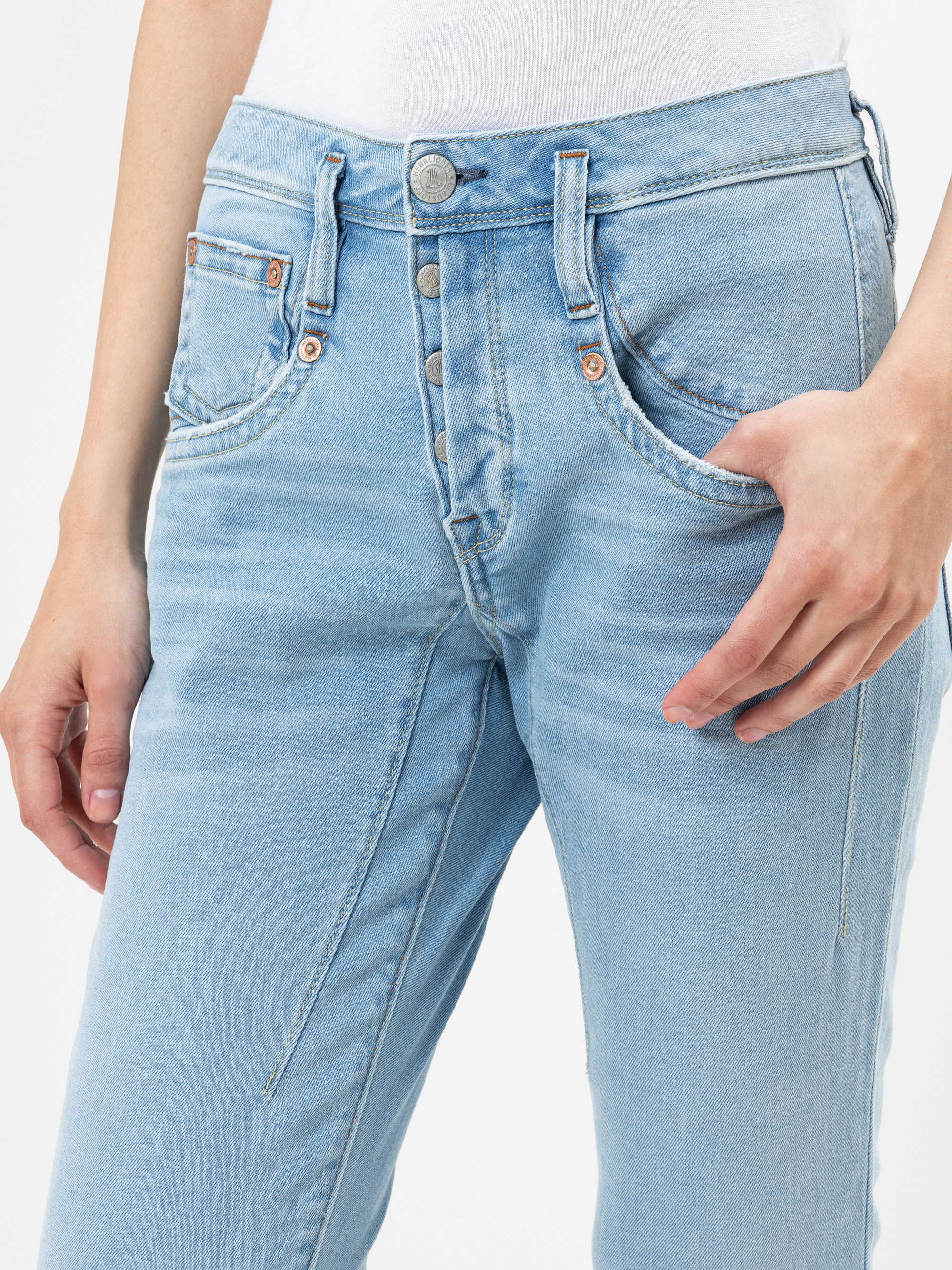 Eine Person, die hellblaue Jeans mit hoher Taille und sichtbaren Knöpfen und ein weißes Oberteil trägt, von der Mitte des Oberkörpers bis zur Mitte des Oberschenkels, wobei eine Hand in der Nähe der Hüfte ruht.