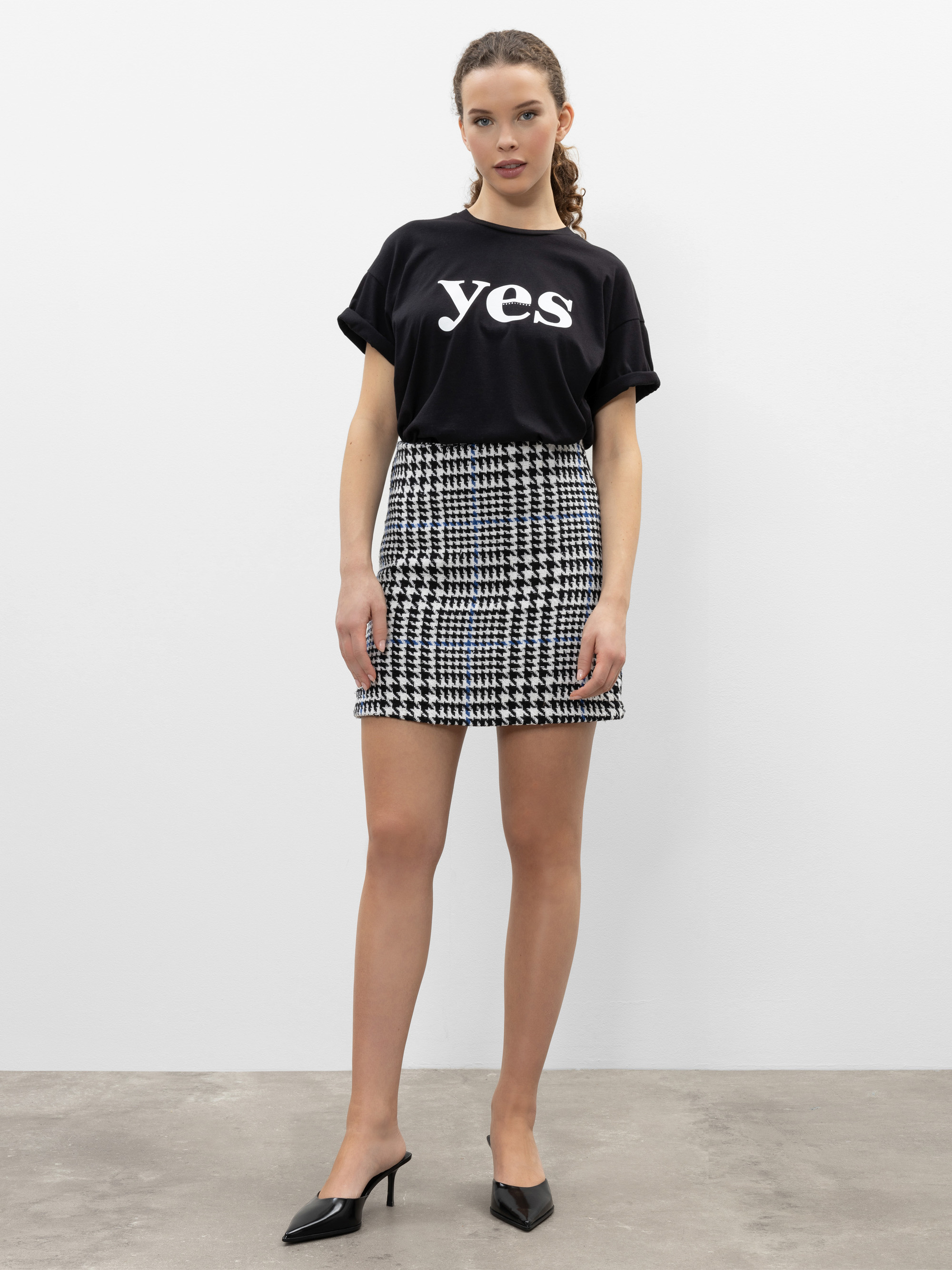 Eine Frau steht vor einer schlichten weißen Wand und trägt ein schwarzes T-Shirt mit dem Wort "Yes" in großen weißen Buchstaben, einen schwarz-weiß karierten Rock und schwarze spitze Absätze.