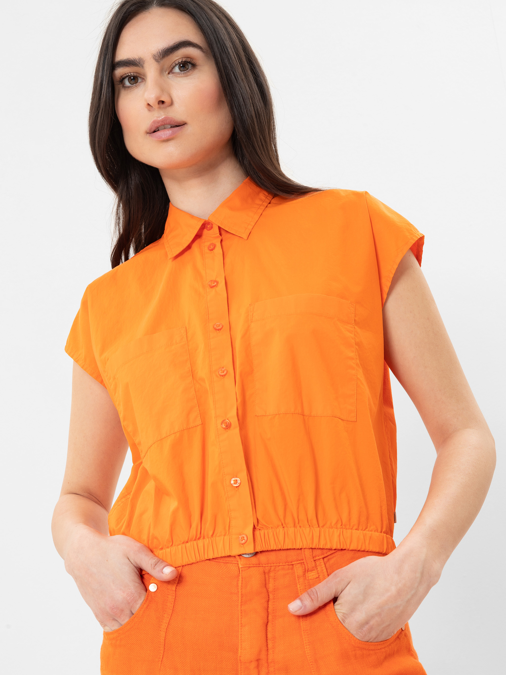 Eine Frau mit langen dunklen Haaren trägt ein leuchtend orangefarbenes, kurzärmeliges Button-up-Hemd und eine dazu passende Hose mit hoher Taille vor einem schlichten weißen Hintergrund.
