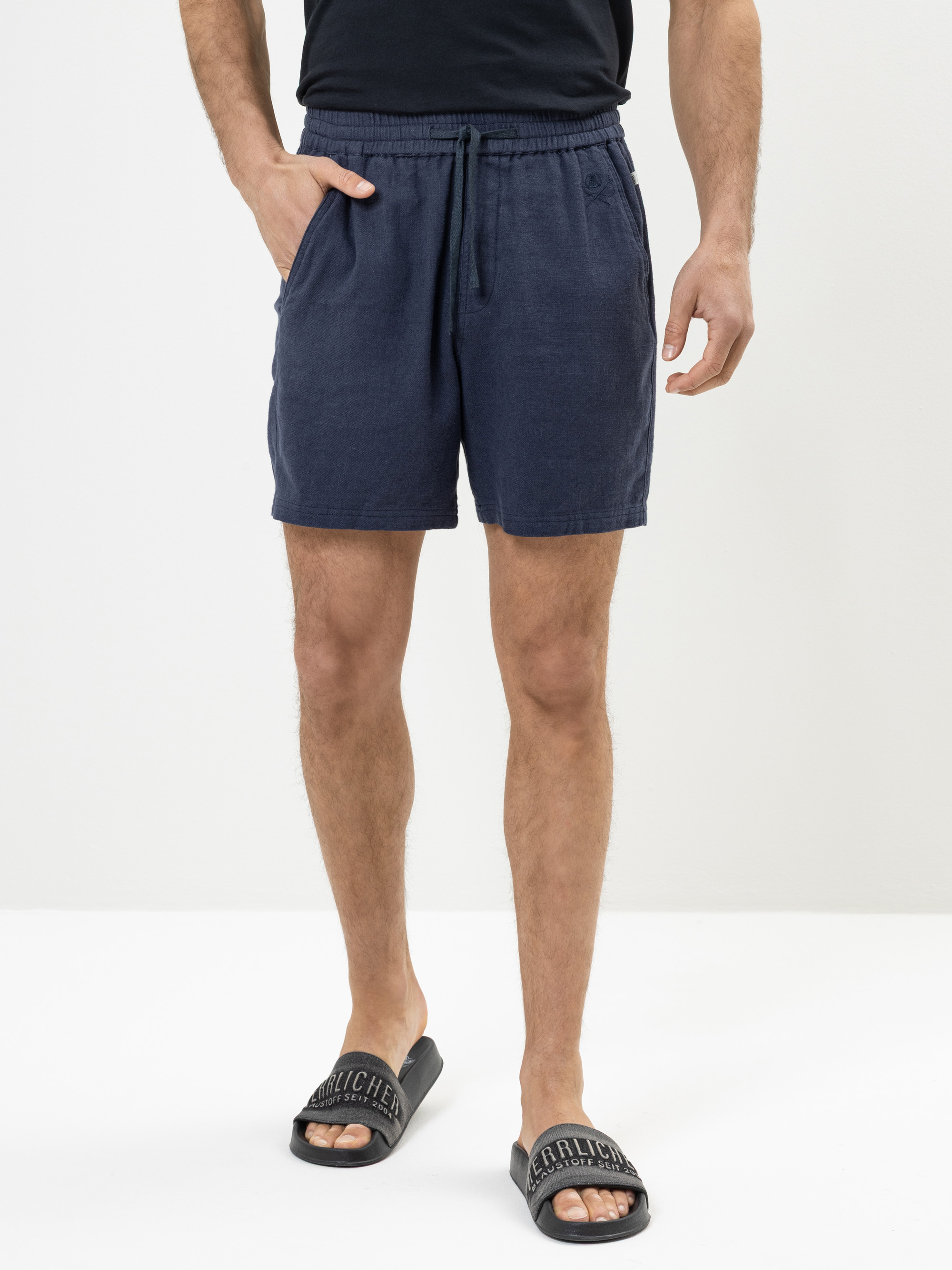 Eine Person, die marineblaue Shorts mit Kordelzug, ein schwarzes T-Shirt und schwarze Slide-Sandalen trägt, steht vor einem schlichten weißen Hintergrund. Ihre Hände stecken in den Taschen der Shorts.