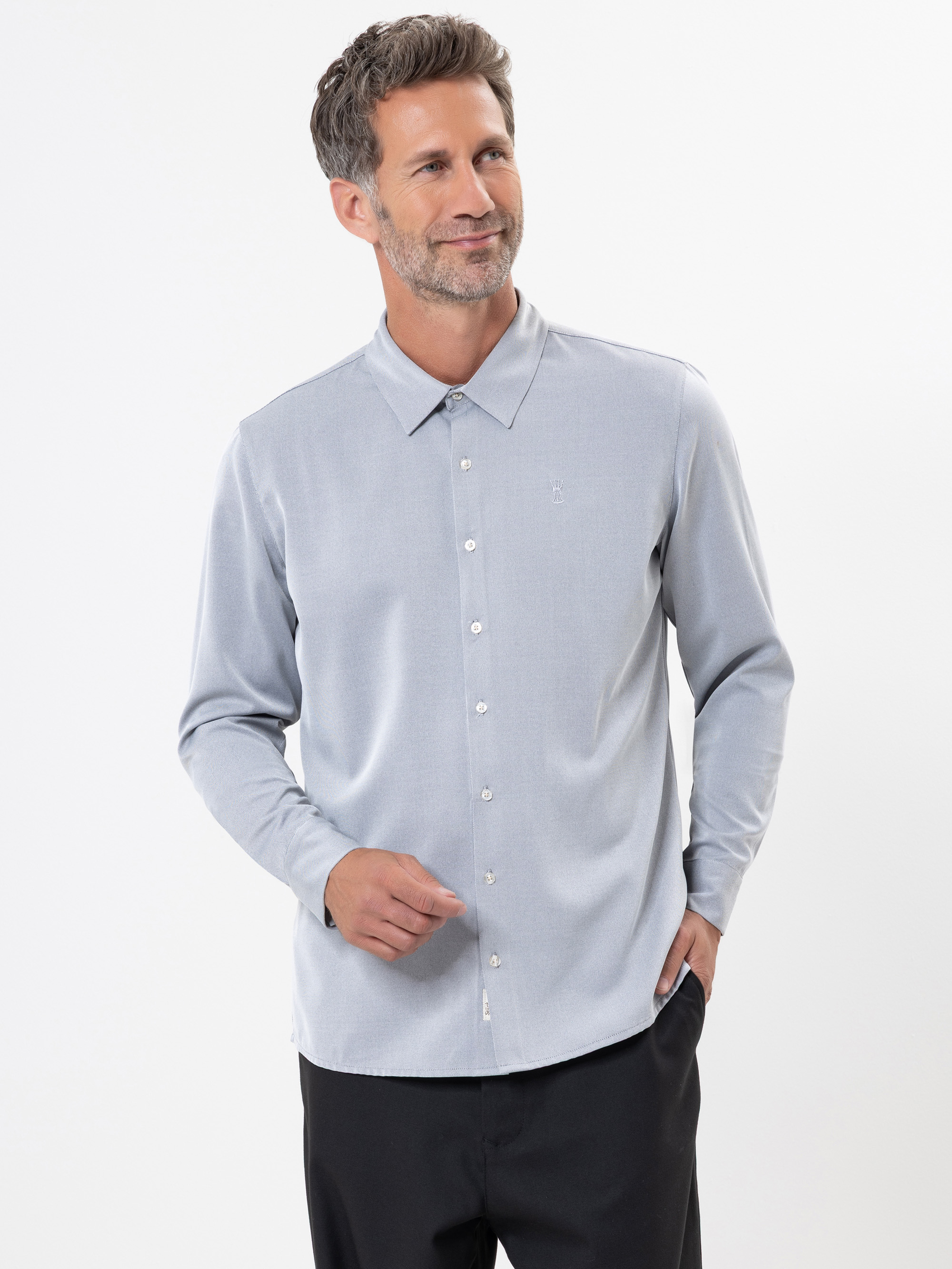 Ein Mann mit kurzem, grauem Haar und gestutztem Bart trägt ein hellgraues Button-up-Hemd und eine schwarze Hose. Er steht vor einem schlichten weißen Hintergrund, lächelt leicht und blickt nach links.