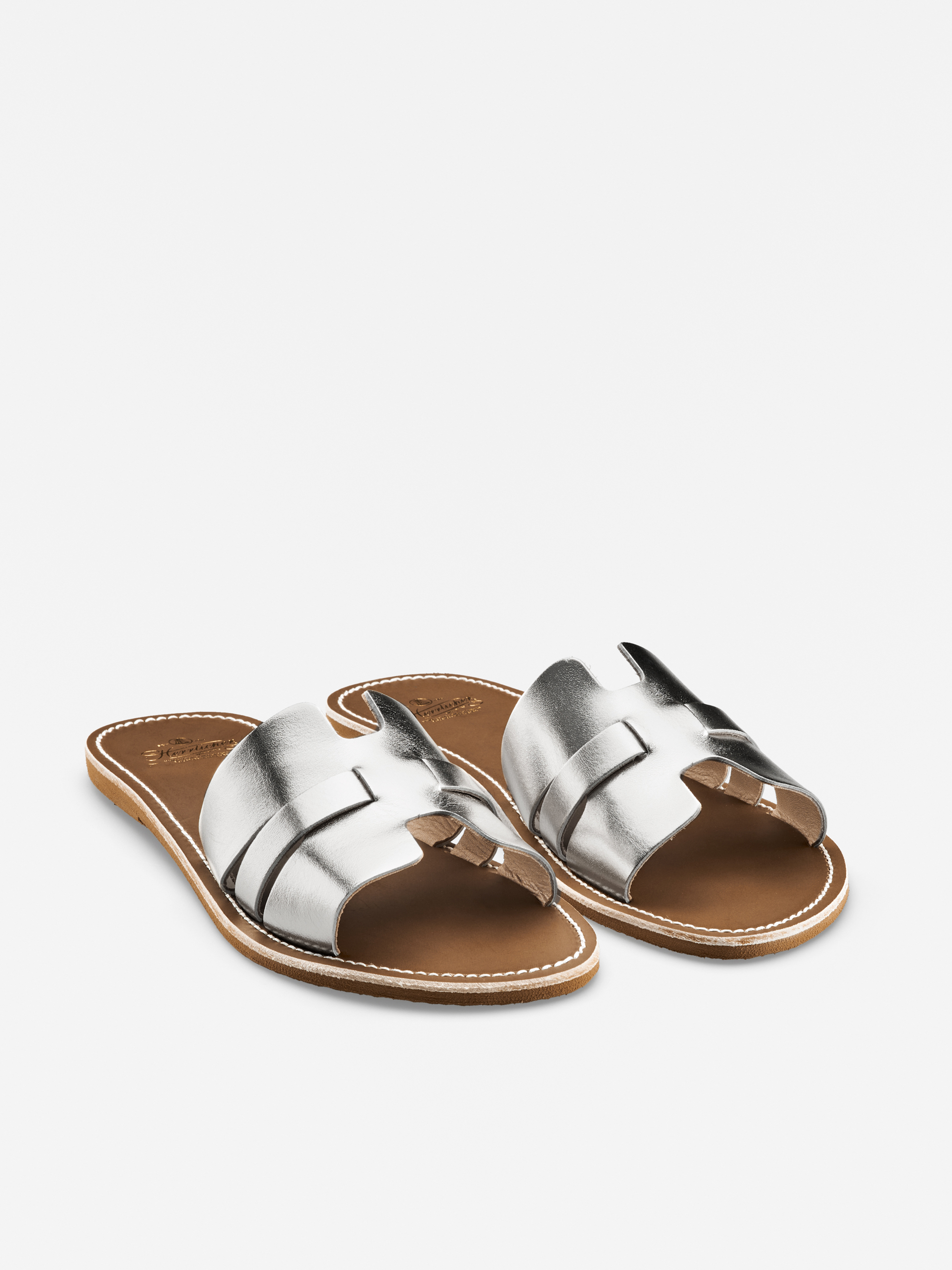 Ein Paar silberne Ledersandalen mit flacher, hellbrauner Sohle und weißen Nähten, mit breiten, sich kreuzenden Riemen und offener Spitze auf weißem Hintergrund.