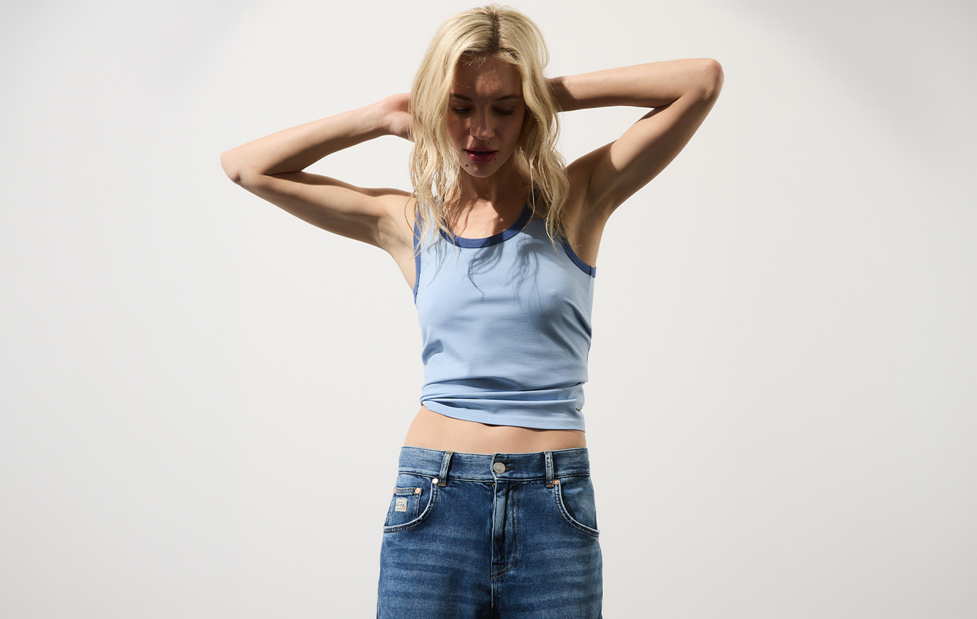 Eine Frau mit langen blonden Haaren trägt ein hellblaues Trägertop und eine blaue Jeans. Sie steht vor einem schlichten weißen Hintergrund, die Arme erhoben und die Hände am Hinterkopf verschränkt.