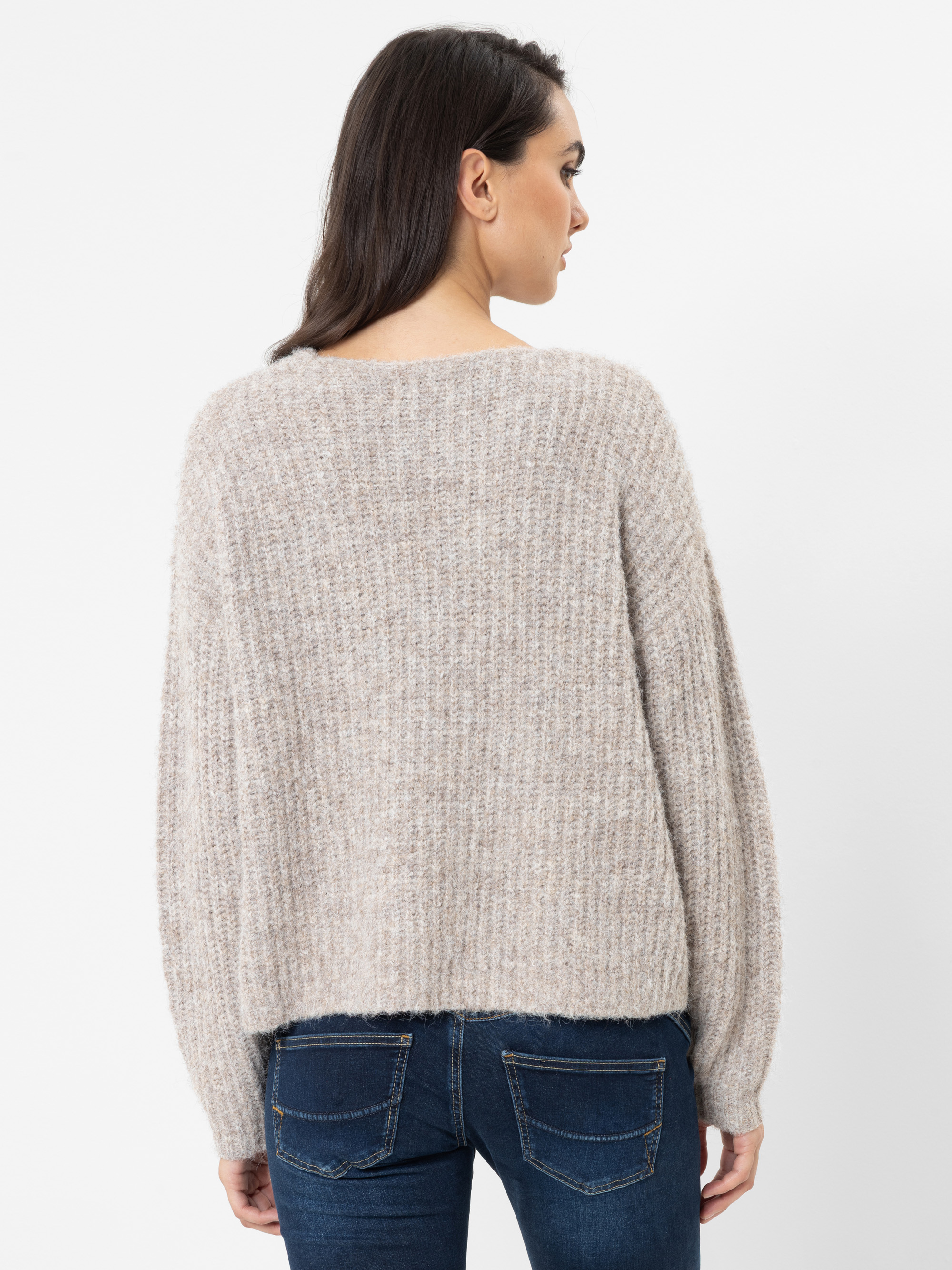 Vor einem schlichten weißen Hintergrund ist eine Frau mit langen dunklen Haaren von hinten zu sehen, die einen lockeren, beigen Strickpullover und eine blaue Jeans trägt.