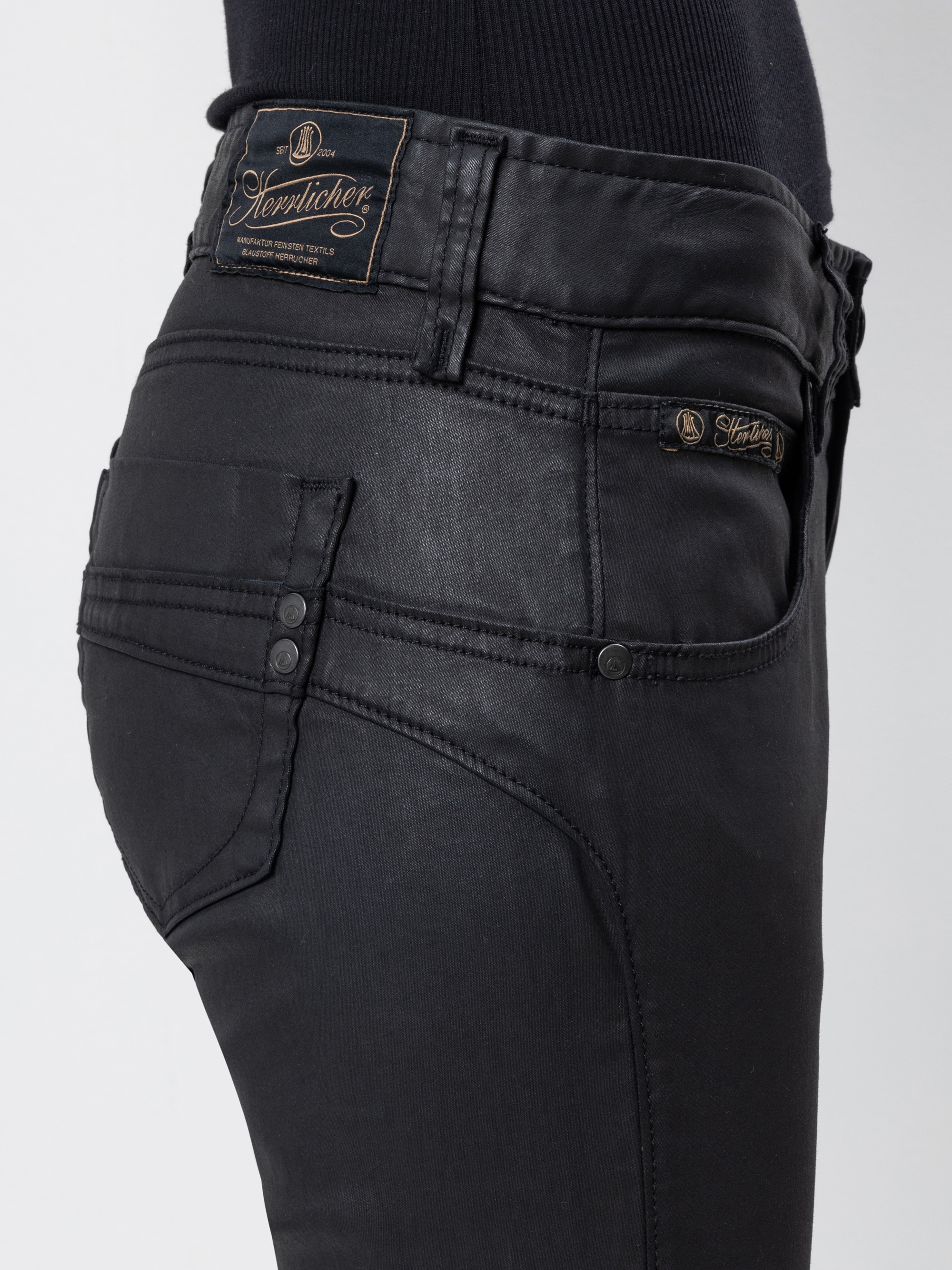 Seitenansicht einer Person, die eine schwarze, taillierte Jeans mit einem Logoaufnäher am Bund und an der Tasche trägt, gepaart mit einem schwarzen Oberteil. Die Jeans hat eine leicht glänzende Textur und Knopfdetails an den Taschen.