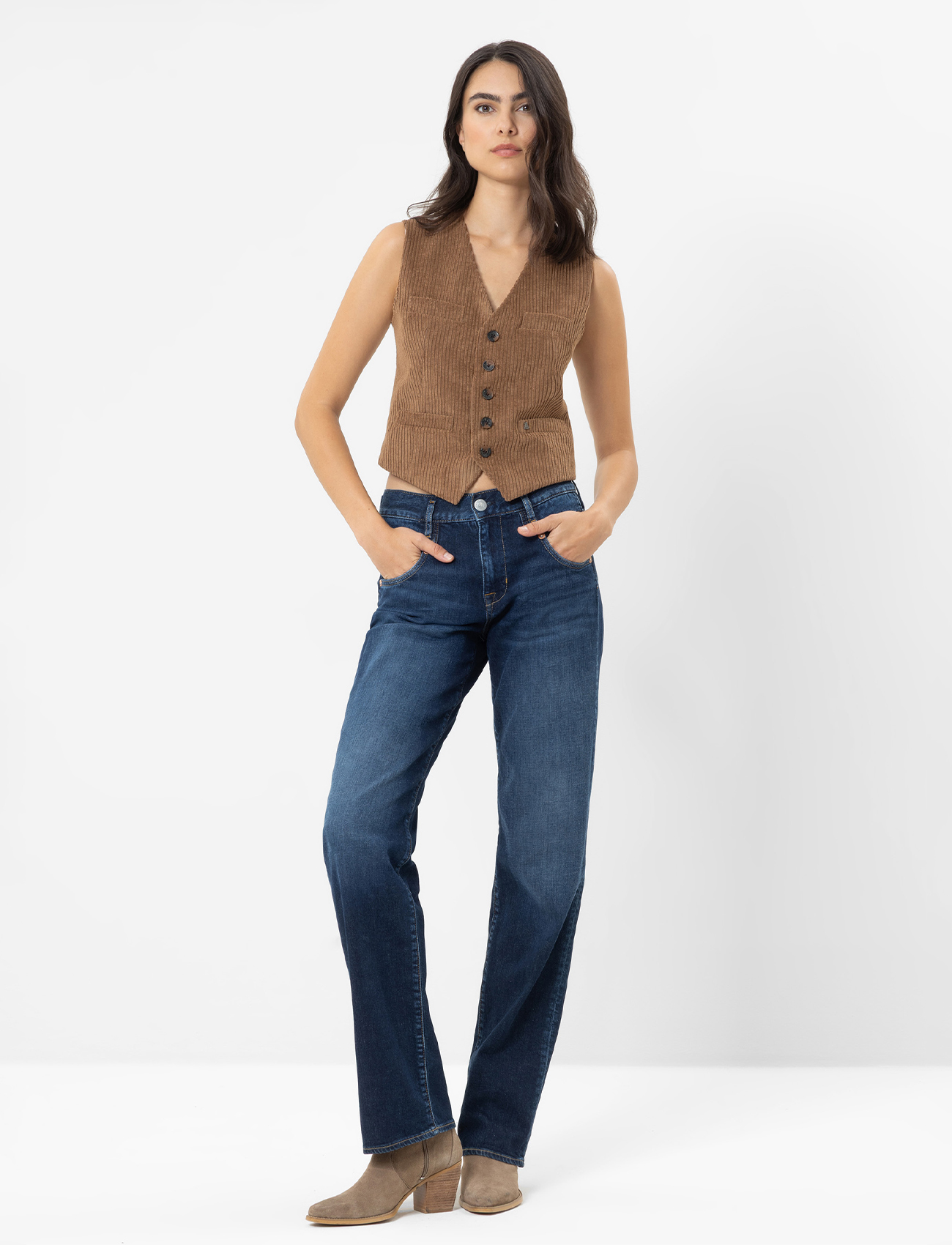 Eine Frau steht vor einem schlichten weißen Hintergrund, trägt ein weißes langärmeliges Oberteil, blaue Jeans und weiße Turnschuhe und hat die Hände in den Taschen ihrer Jeans.