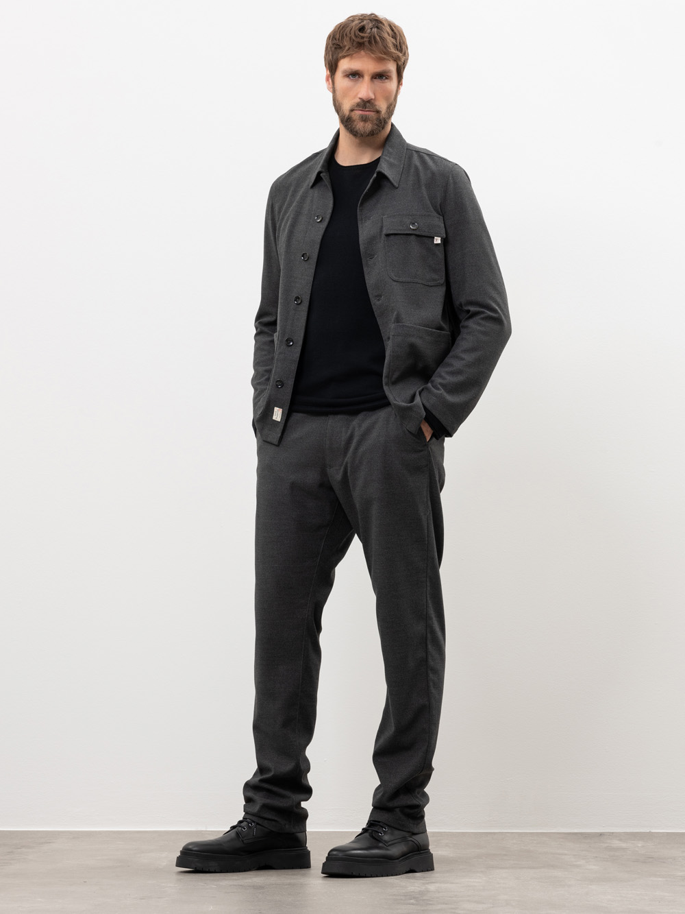 Ein Mann steht vor einer schlichten weißen Wand, trägt eine dunkelgraue Jacke mit Knöpfen, eine dazu passende Hose, ein schwarzes Hemd und schwarze Schuhe, hat die Hände in den Taschen und schaut in die Kamera.