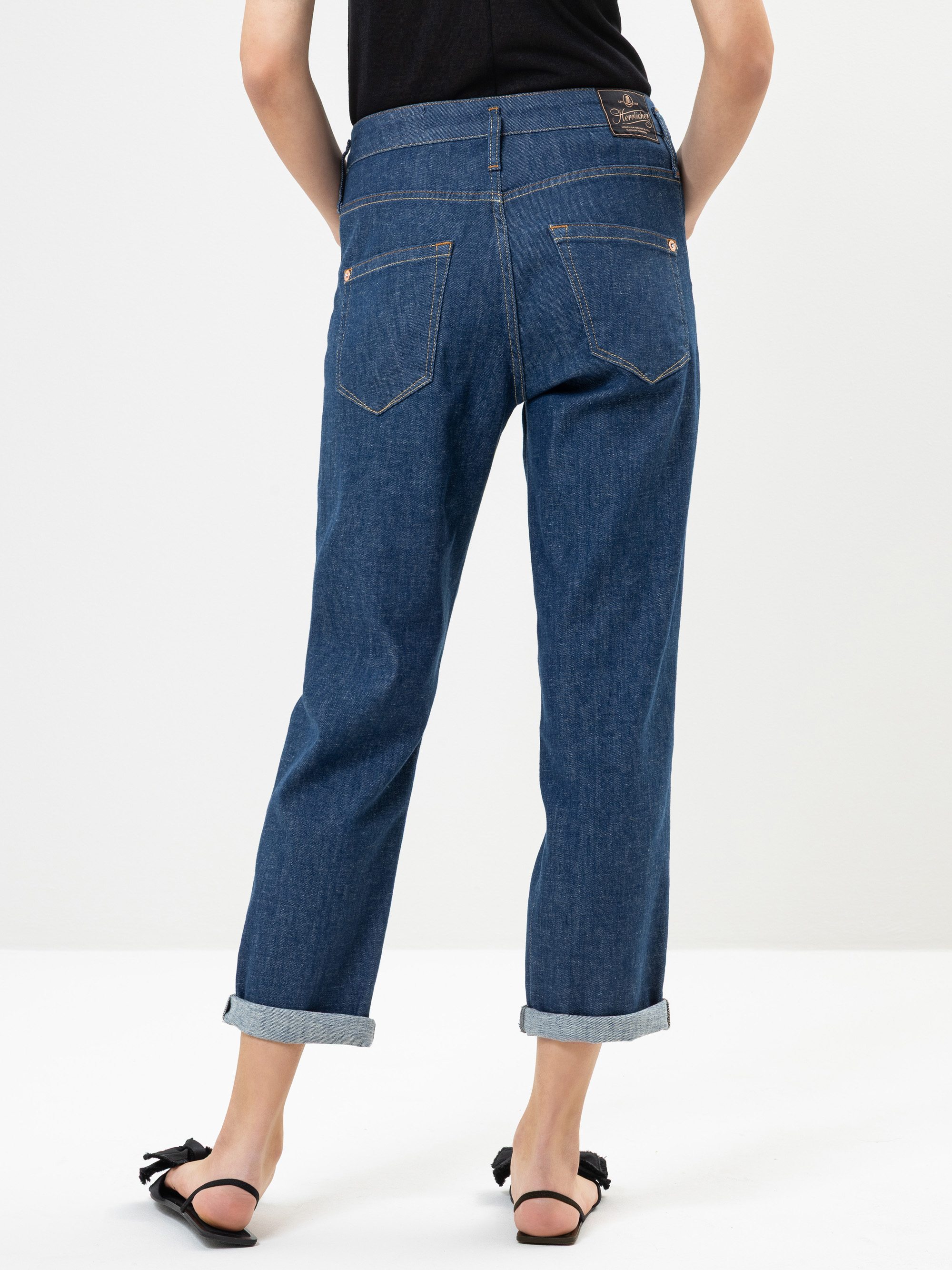Eine Person, die mit dem Rücken zur Kamera steht, trägt eine blaue Jeans mit Manschetten und schwarze Sandalen mit Schleifen; ihre Hände ruhen auf den Hüften, und der Hintergrund ist schlicht weiß.
