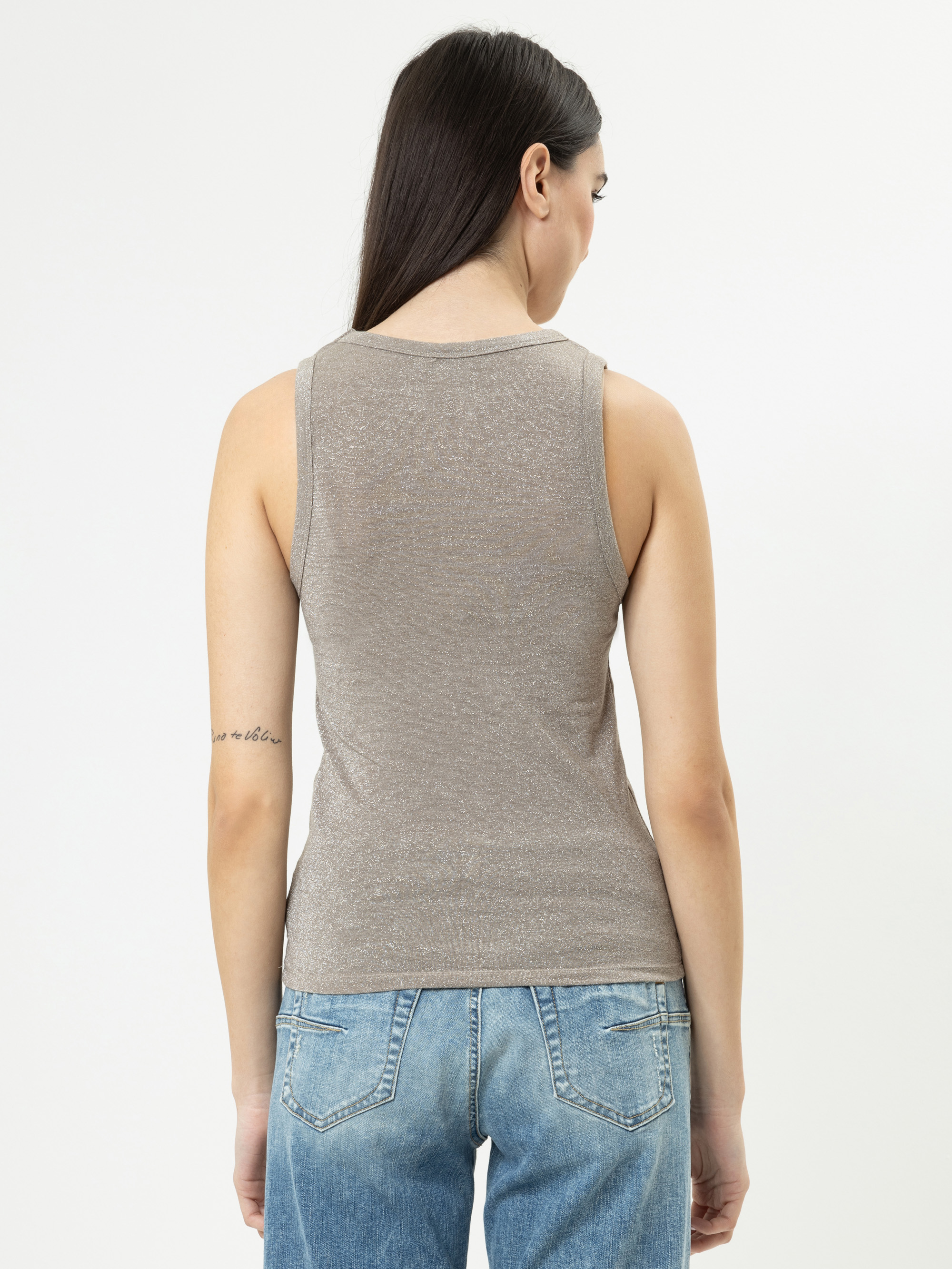 Eine Frau mit langen dunklen Haaren, die ein ärmelloses graues Oberteil und blaue Jeans trägt, steht mit dem Rücken zur Kamera vor einem schlichten weißen Hintergrund. Auf ihrem linken Oberarm ist eine kleine Tätowierung zu sehen.