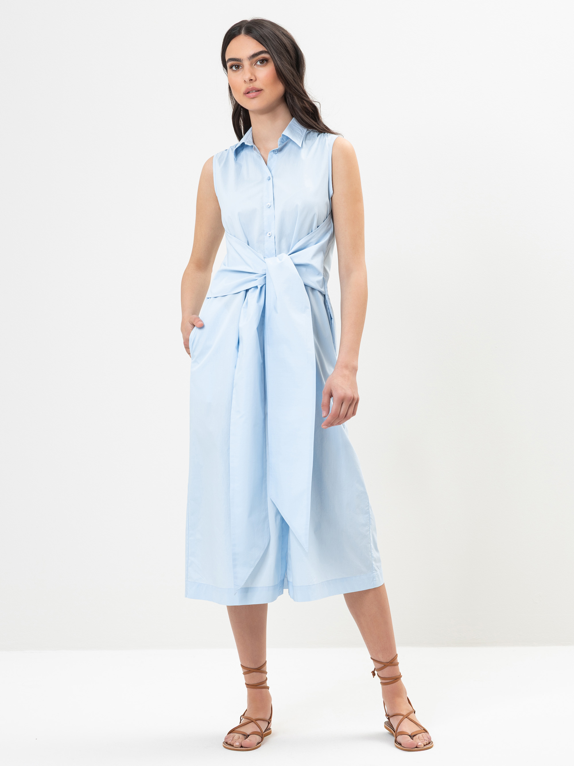 Eine Frau steht vor einem schlichten weißen Hintergrund und trägt ein ärmelloses, hellblaues Midikleid mit Kragen, einer großen Krawatte vorne und Seitentaschen. Sie hat langes dunkles Haar und trägt braune Riemchensandalen.