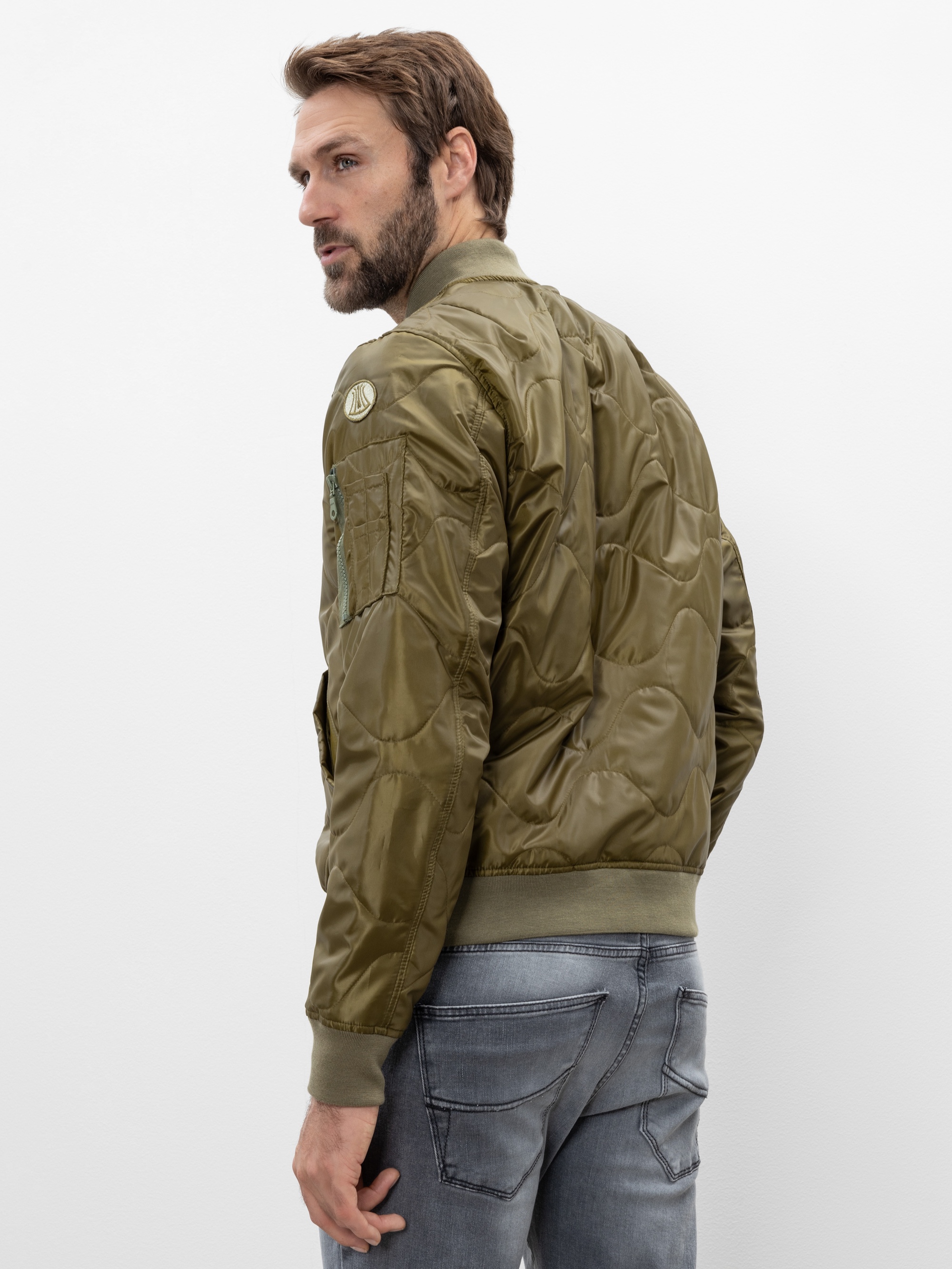 Ein Mann mit hellbraunem Haar und Bart trägt eine olivgrüne Bomberjacke und verwaschene graue Jeans. Er steht mit dem Rücken zur Kamera und blickt über seine linke Schulter vor einem schlichten weißen Hintergrund.