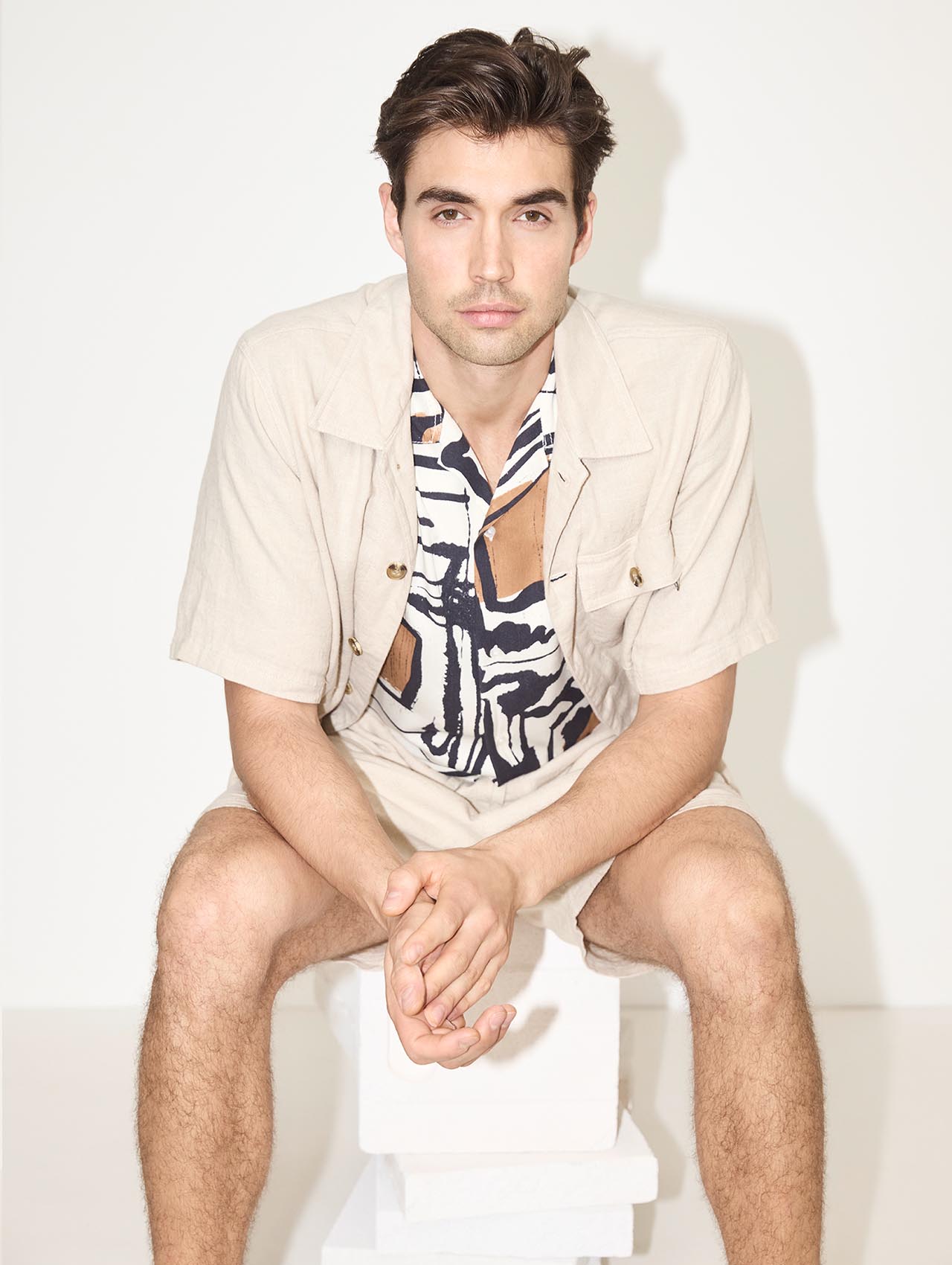 Ein Mann mit dunklem Haar sitzt auf einem weißen Block und trägt ein hellbeiges kurzärmeliges Hemd über einem gemusterten Hemd und dazu passende Shorts. Er blickt mit gefalteten Händen in die Kamera, vor einem schlichten hellen Hintergrund.