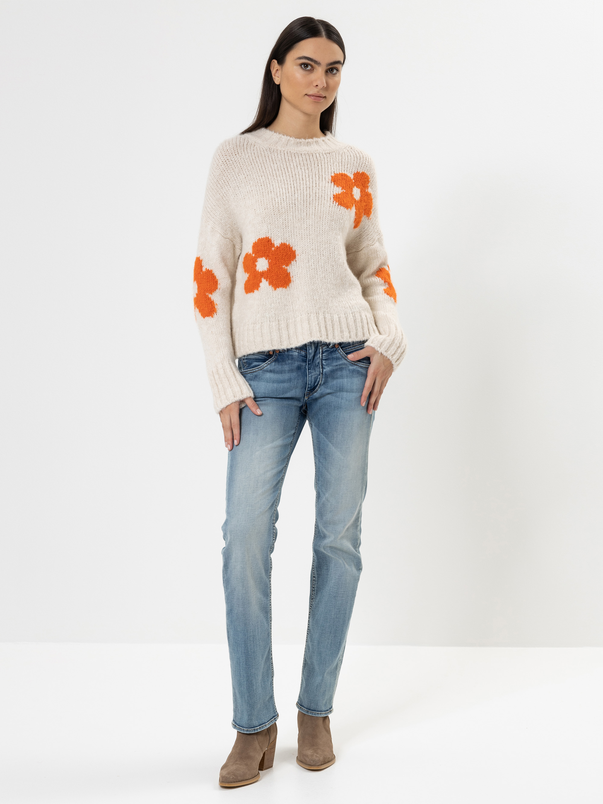 Eine Frau steht vor einem schlichten weißen Hintergrund und trägt einen cremefarbenen Pullover mit orangefarbenen Blumenmustern, blaue Jeans und braune Ankle Boots. Sie hat langes dunkles Haar und einen neutralen Gesichtsausdruck.