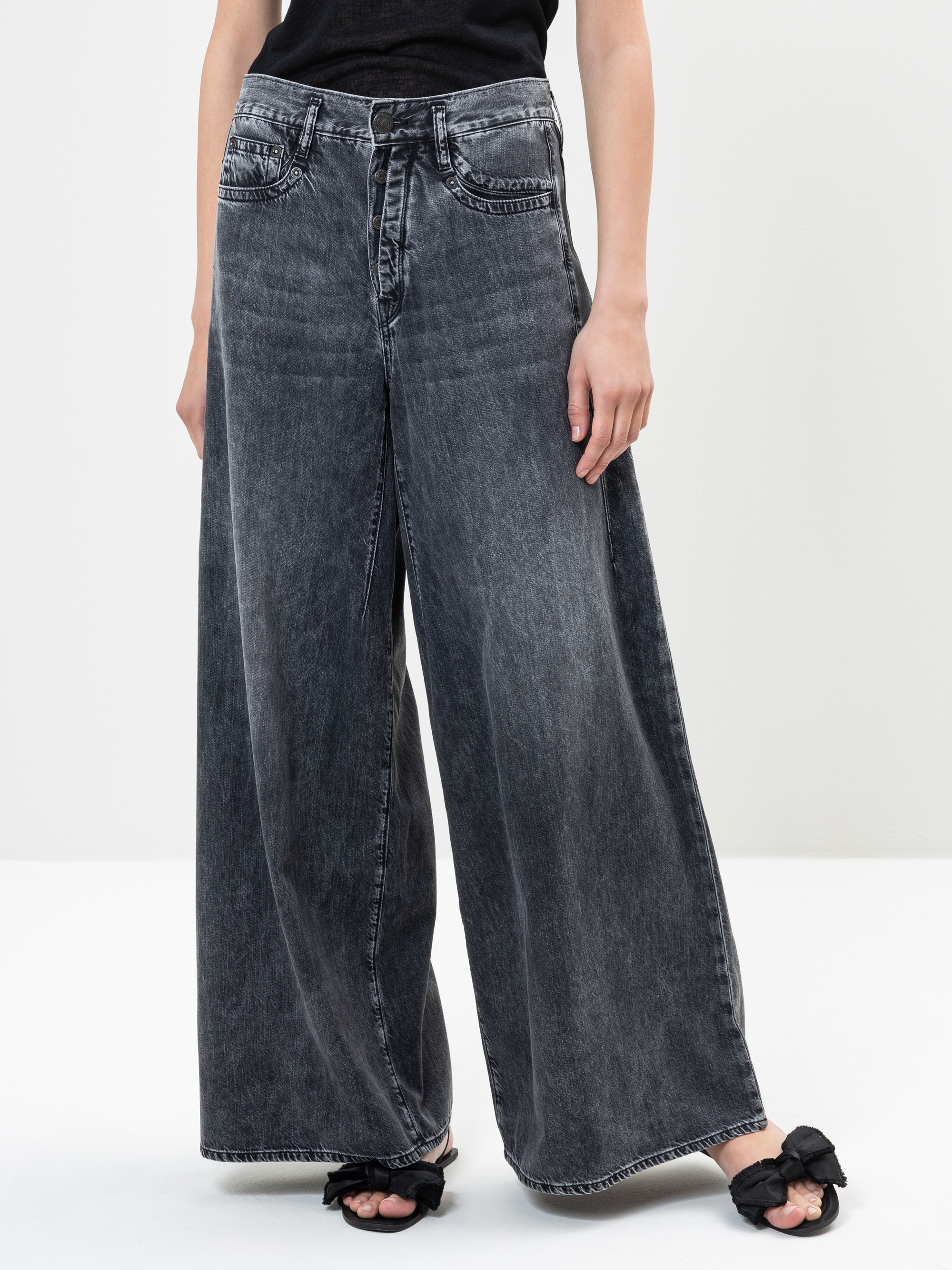 Person, die eine schwarze, gewaschene Denim-Jeans mit hoher Taille und weitem Bein zu einem schwarzen Oberteil trägt. Die Jeans ist bodenlang und wird mit schwarzen Sandalen mit Schleifen gepaart. Der Hintergrund ist einfarbig weiß.