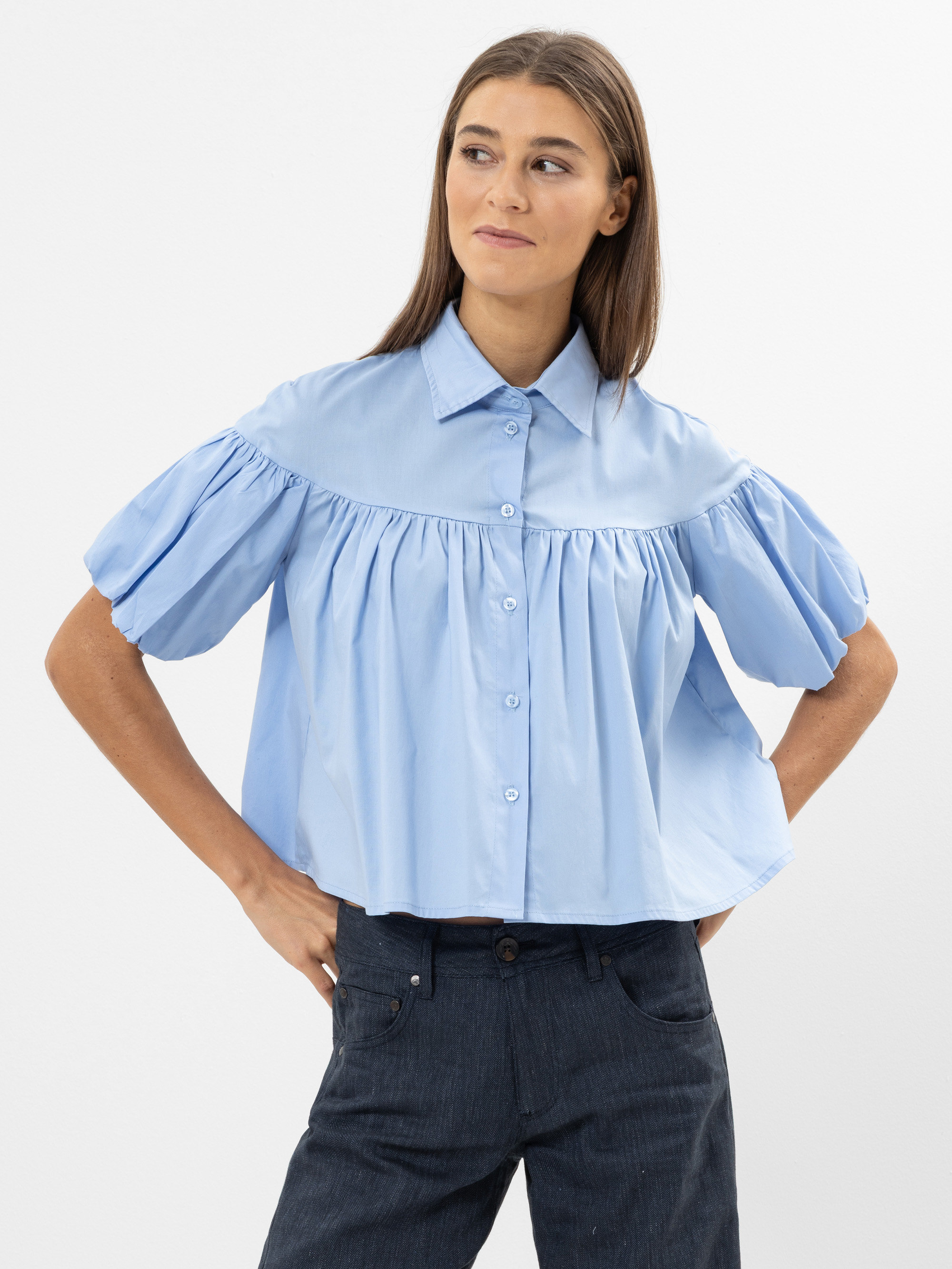 Eine Frau, die eine hellblaue, kurzärmelige, hochgeknöpfte Bluse mit Puffärmeln und eine dunkle Jeans trägt, steht vor einem schlichten weißen Hintergrund, schaut leicht zur Seite und lächelt sanft.