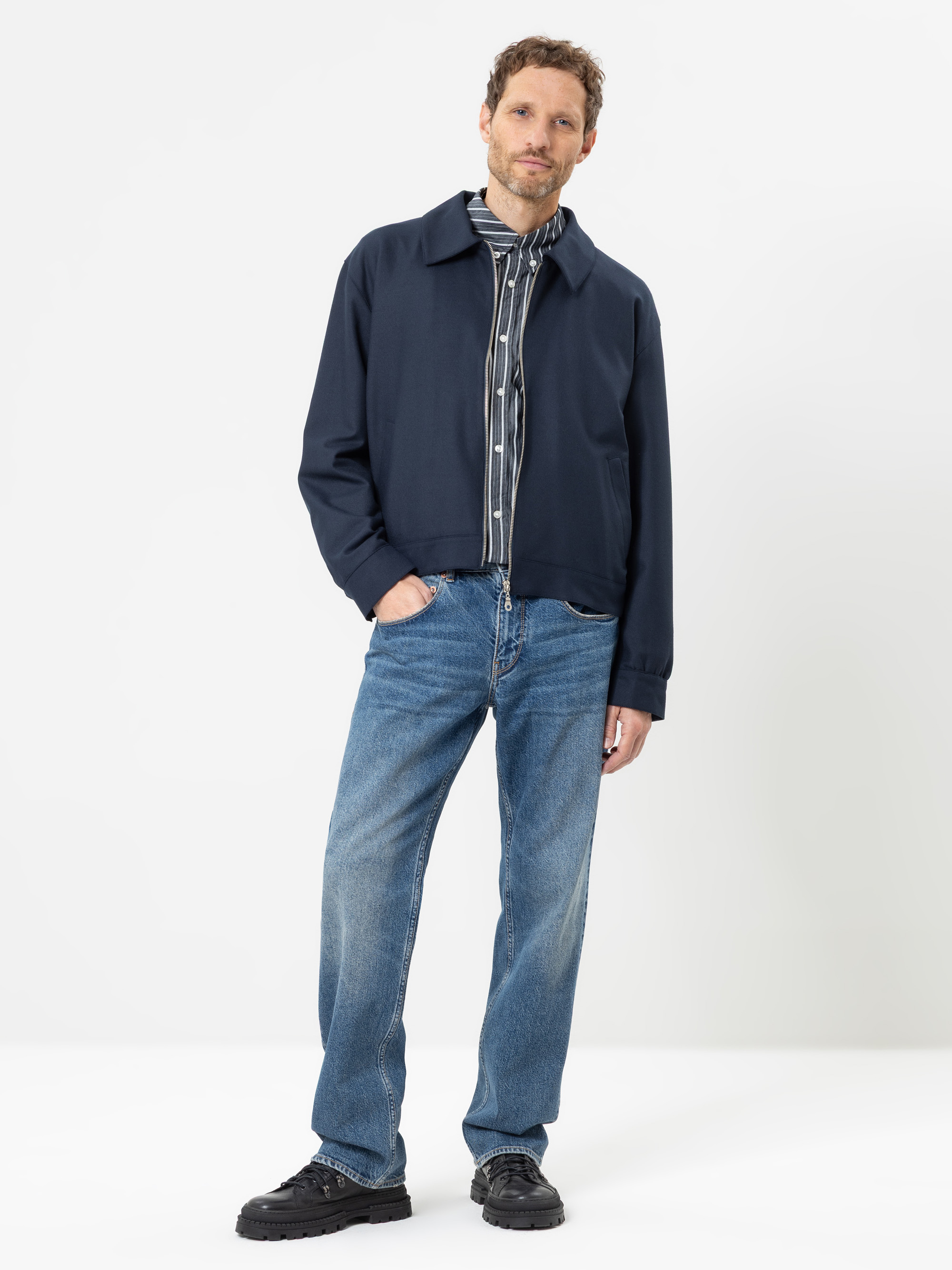 Ein Mann steht vor einem schlichten weißen Hintergrund. Er trägt eine marineblaue Jacke, ein gestreiftes Hemd mit Knöpfen, blaue Jeans und schwarze Schuhe. Eine Hand steckt in seiner Hosentasche, und er wirkt ganz entspannt.