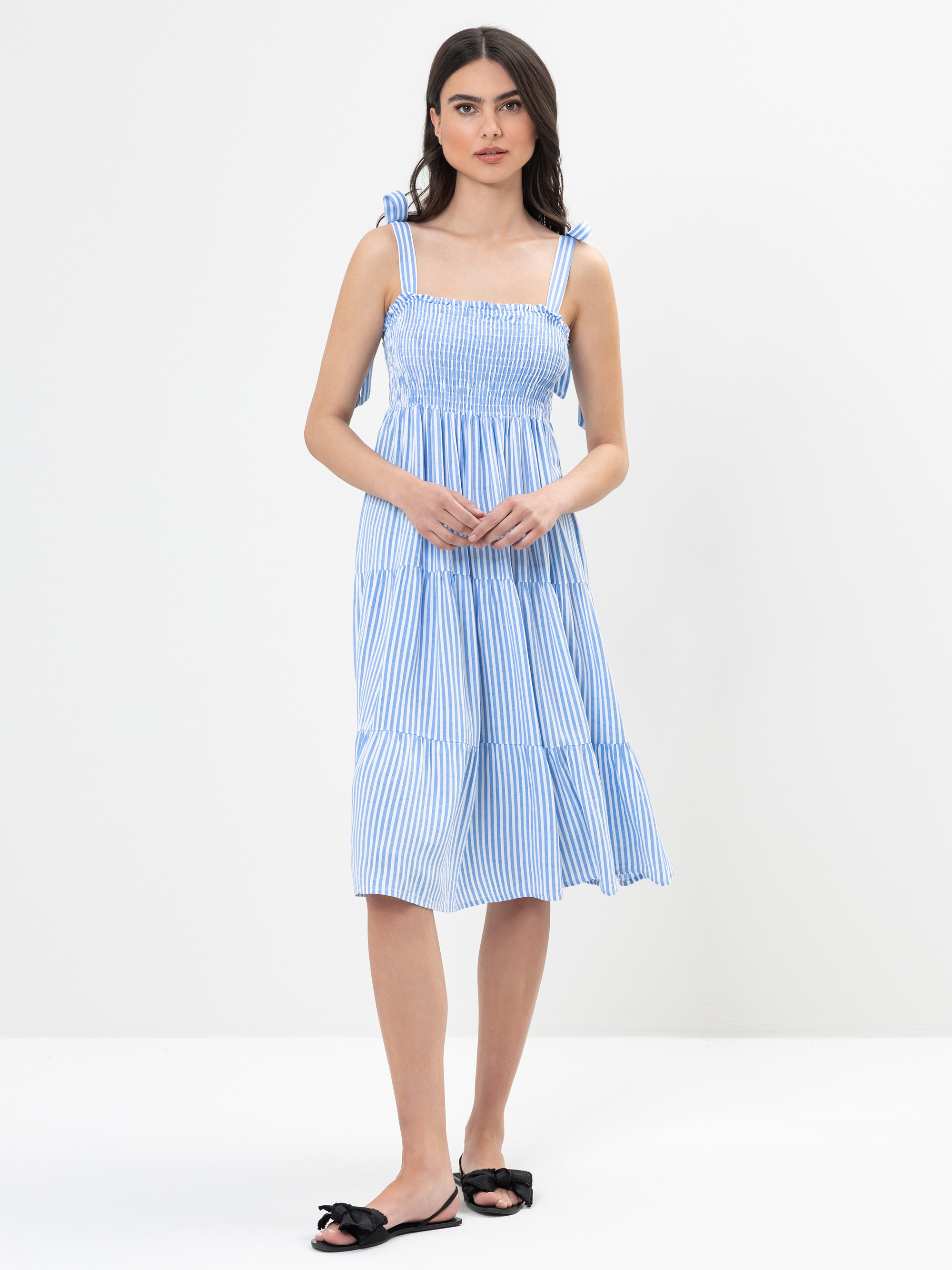 Eine Frau in einem blau-weiß vertikal gestreiften ärmellosen Kleid mit Bindebändern steht vor einem schlichten weißen Hintergrund. Sie hat langes dunkles Haar und trägt schwarze flache Sandalen.
