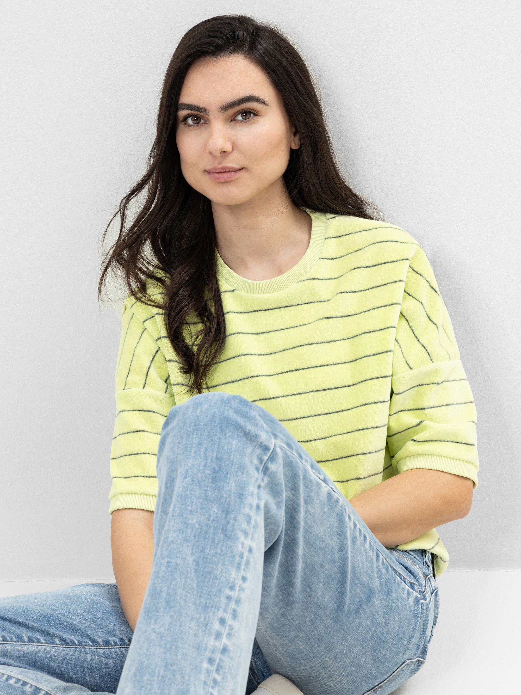 Eine Person mit langen dunklen Haaren sitzt an einer hellen Wand, trägt einen hellgelb gestreiften Pullover und hellblaue Jeans und schaut mit neutralem Blick in die Kamera.