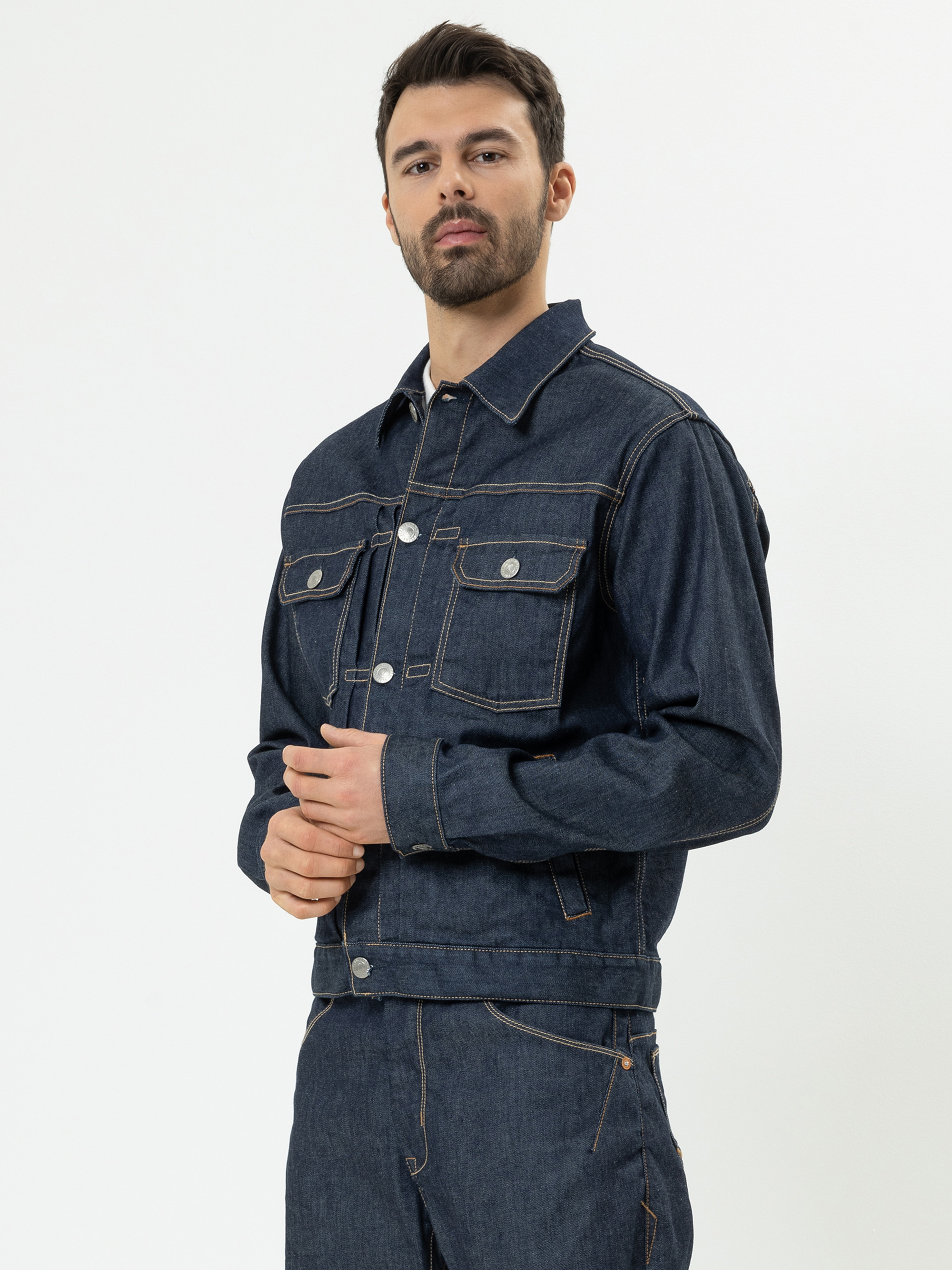 Ein Mann mit dunklem Haar und Bart trägt eine dunkelblaue Jeansjacke und dazu passende Jeans. Er steht leicht zur Seite gewandt, die Hände vor sich verschränkt, vor einem schlichten weißen Hintergrund.