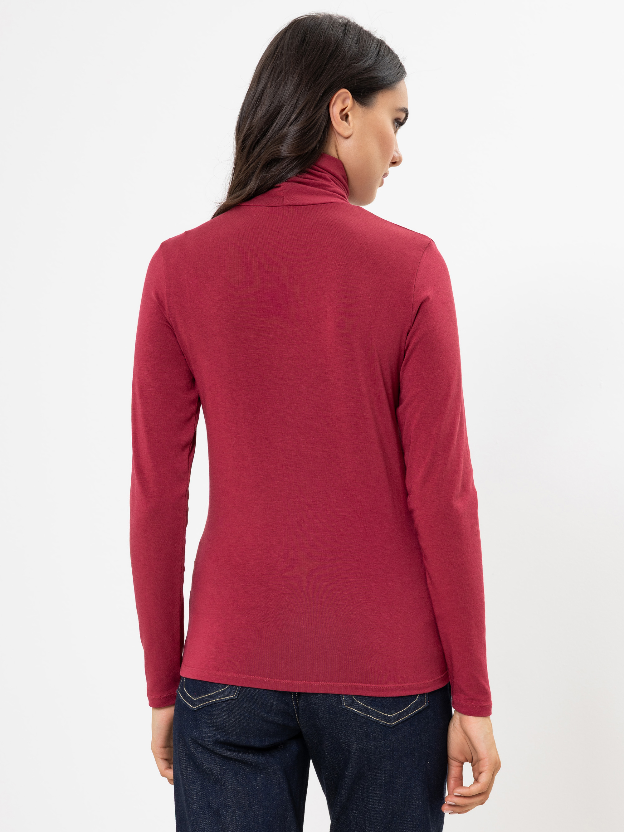 Eine Frau mit langen dunklen Haaren, die ein langärmeliges, rotes Rollkragenoberteil und dunkle Jeans trägt, wird von hinten vor einem schlichten weißen Hintergrund gezeigt.