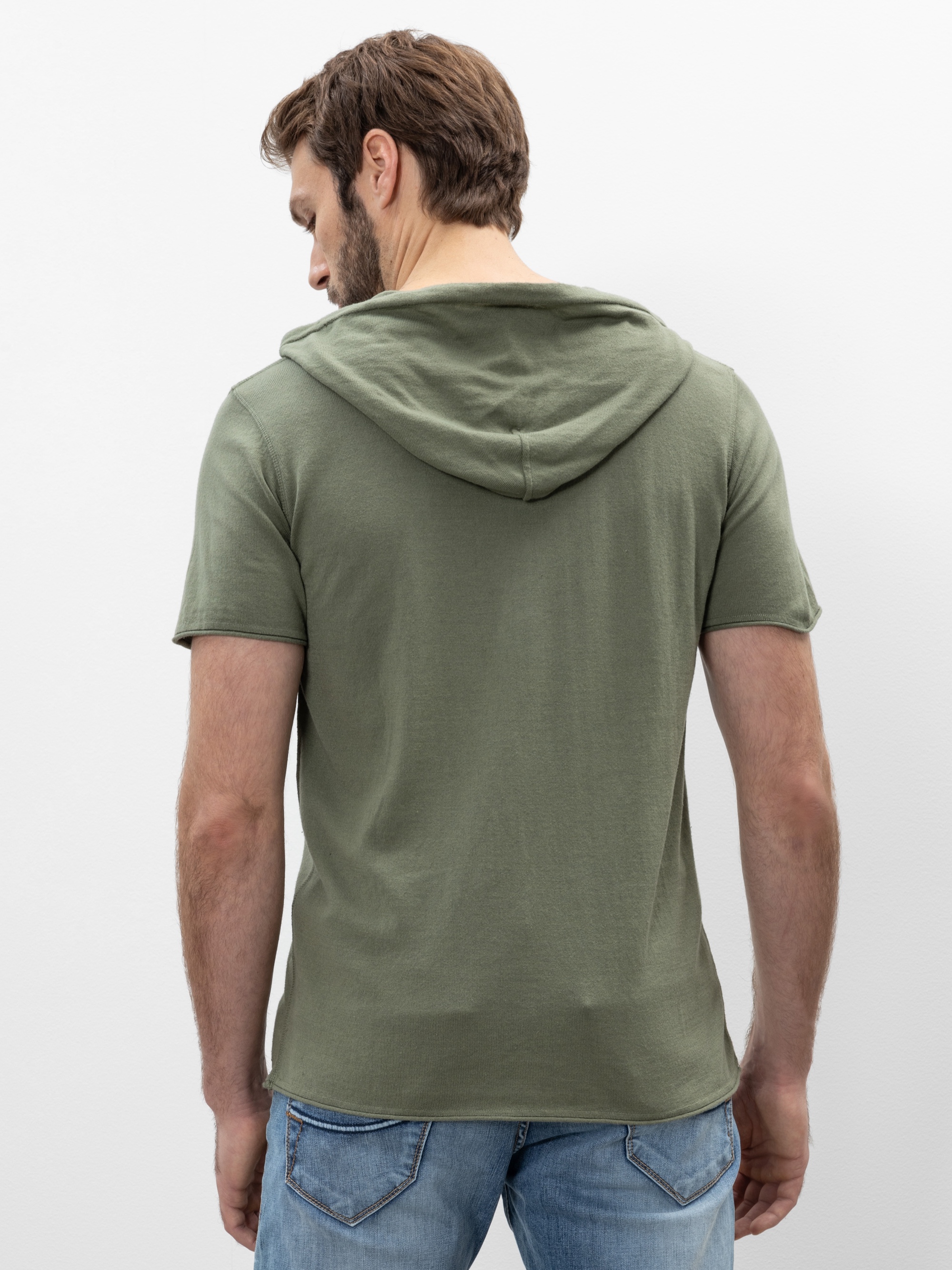 Ein Mann mit hellbraunem Haar steht mit dem Rücken zur Kamera, trägt ein kurzärmeliges, olivgrünes Kapuzen-T-Shirt und blaue Jeans vor einem schlichten weißen Hintergrund.