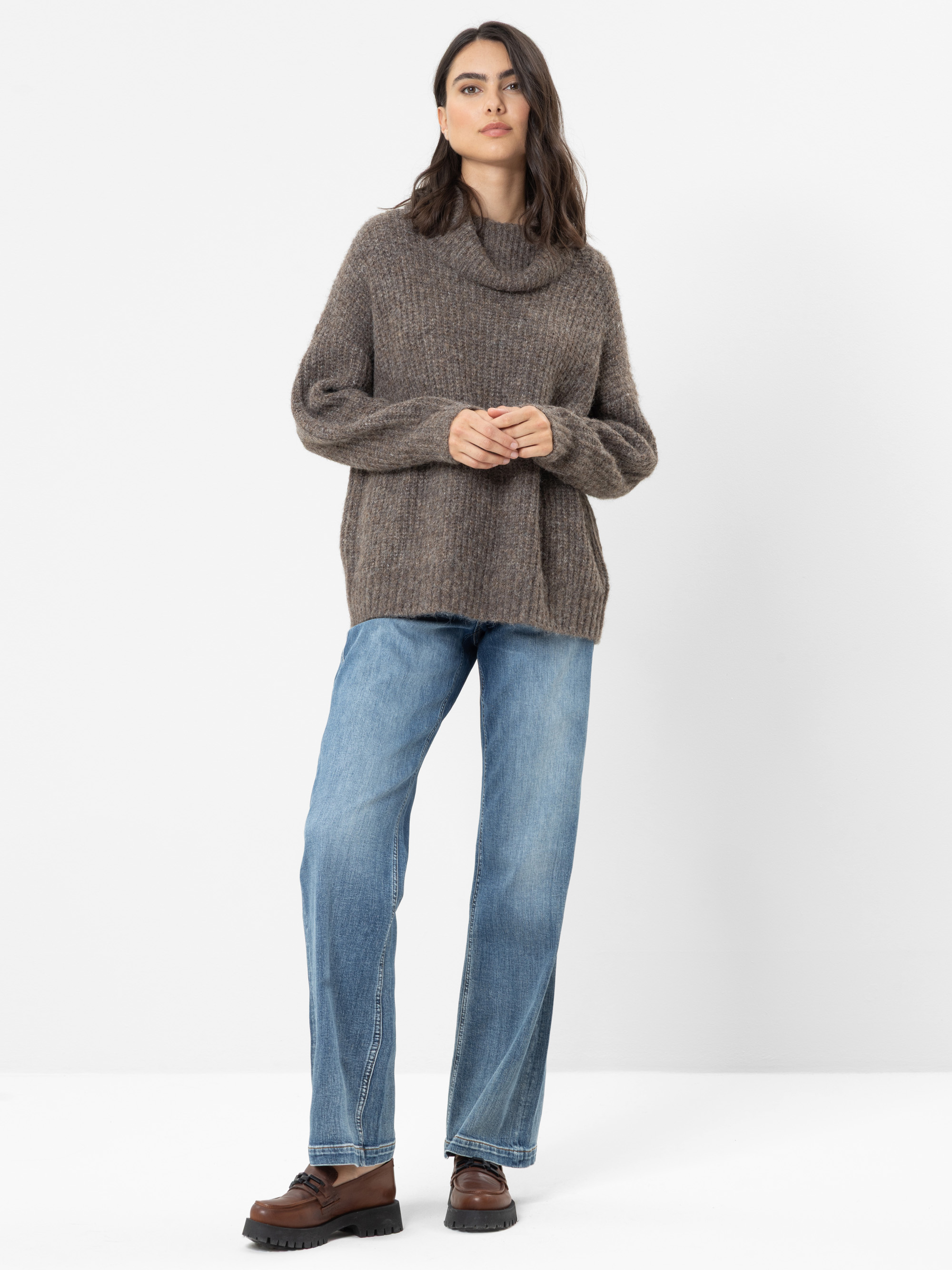 Eine Frau steht vor einem weißen Hintergrund und trägt einen lockeren braunen Strickpullover, blaue Jeans und braune Slipper. Sie hat langes dunkles Haar und einen ausdruckslosen Gesichtsausdruck; ihre Hände liegen entspannt vor ihrem Körper.