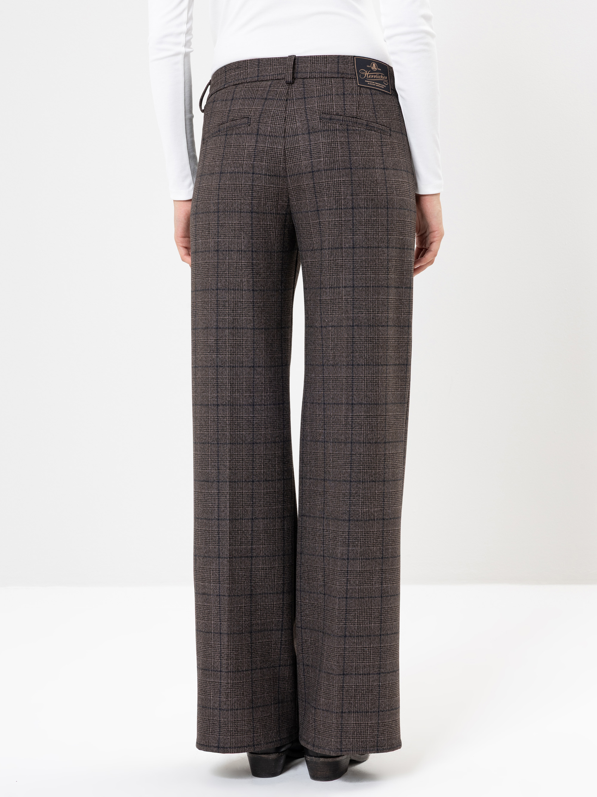 Eine Person, die eine hochtaillierte, weit geschnittene karierte Hose in Braun und Schwarz trägt, steht mit dem Gesicht zur Seite und hat die Arme locker an der Seite. Sie trägt außerdem ein weißes langärmeliges Oberteil und schwarze Schuhe.