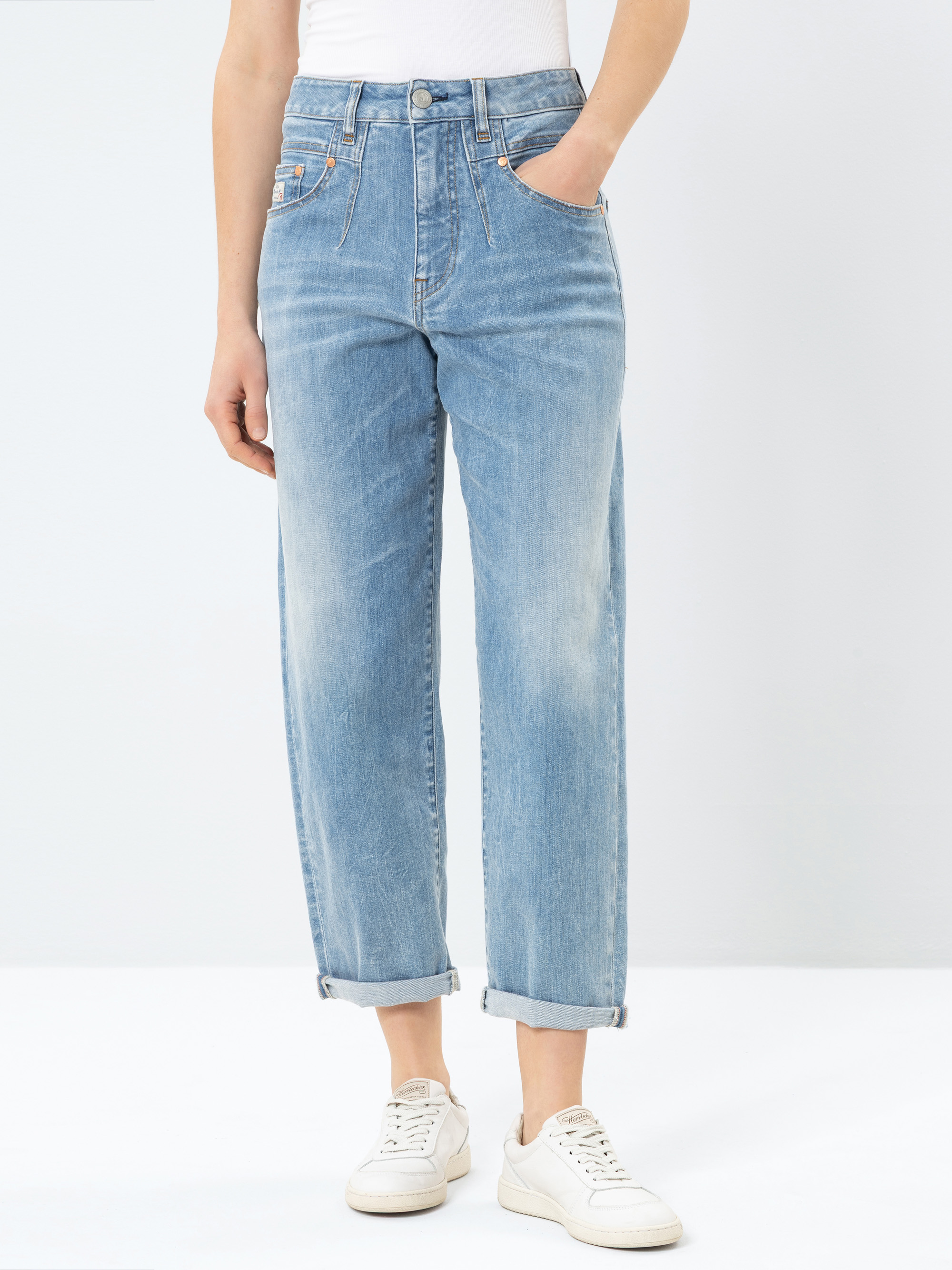 Eine Person, die eine hellblaue Jeans mit hoher Taille und geradem Bein und aufgerollten Manschetten, ein weißes Oberteil und weiße Turnschuhe trägt, steht vor einem schlichten weißen Hintergrund. Die Hände sind in den Hosentaschen der Person.