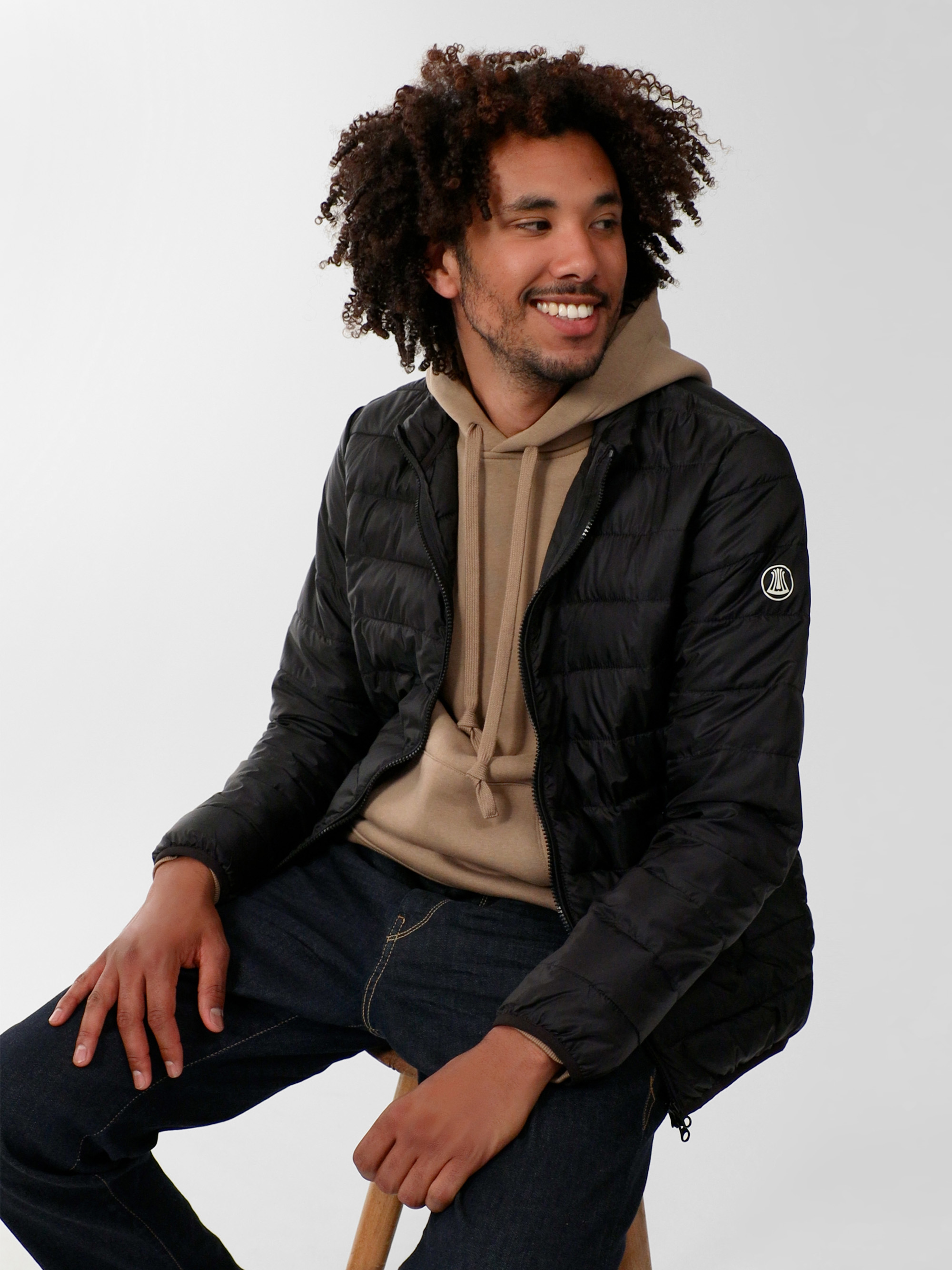 Ein Mann mit lockigem Haar, der einen beigen Kapuzenpulli unter einer schwarzen Kapuzenjacke und dunkle Jeans trägt, sitzt auf einem Holzhocker und blickt vor einem schlichten, hellen Hintergrund lächelnd leicht zur Seite.