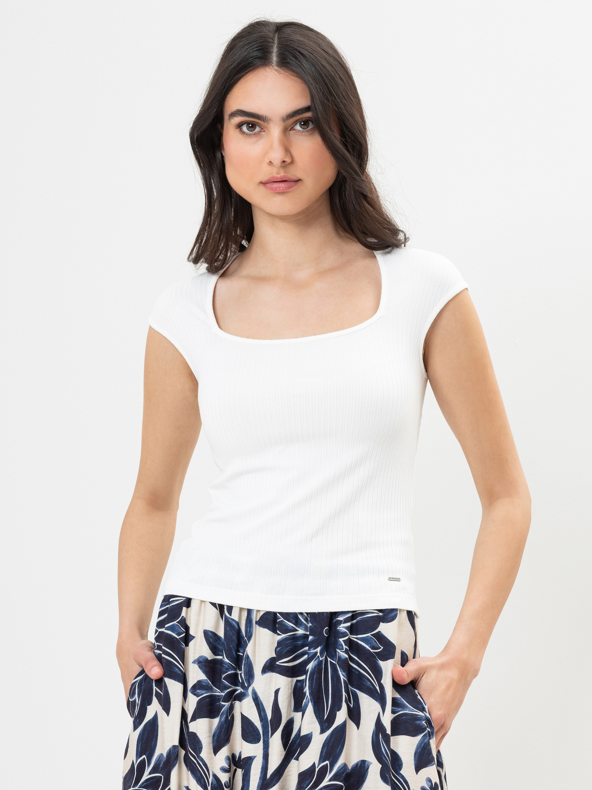 Eine Frau mit langen, dunklen Haaren trägt ein weißes, kurzärmeliges Oberteil mit quadratischem Ausschnitt und einen Rock mit blauem Blumenmuster. Sie steht vor einem schlichten weißen Hintergrund und hat die Hände in den Rocktaschen.