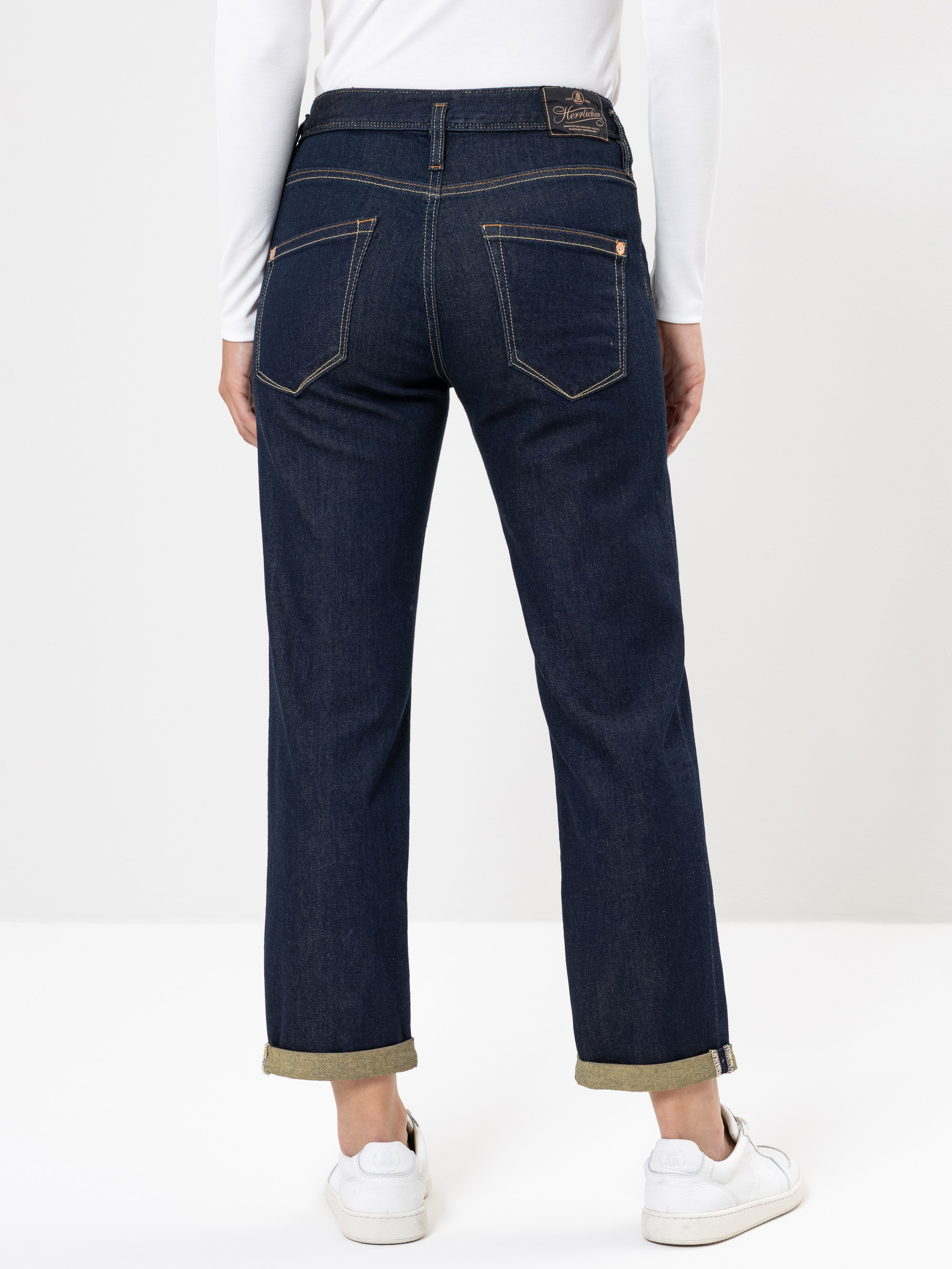 Person, die dunkelblaue Jeans mit Bündchen, ein weißes langärmeliges Oberteil und weiße Turnschuhe trägt, von hinten vor einem schlichten hellen Hintergrund dargestellt.