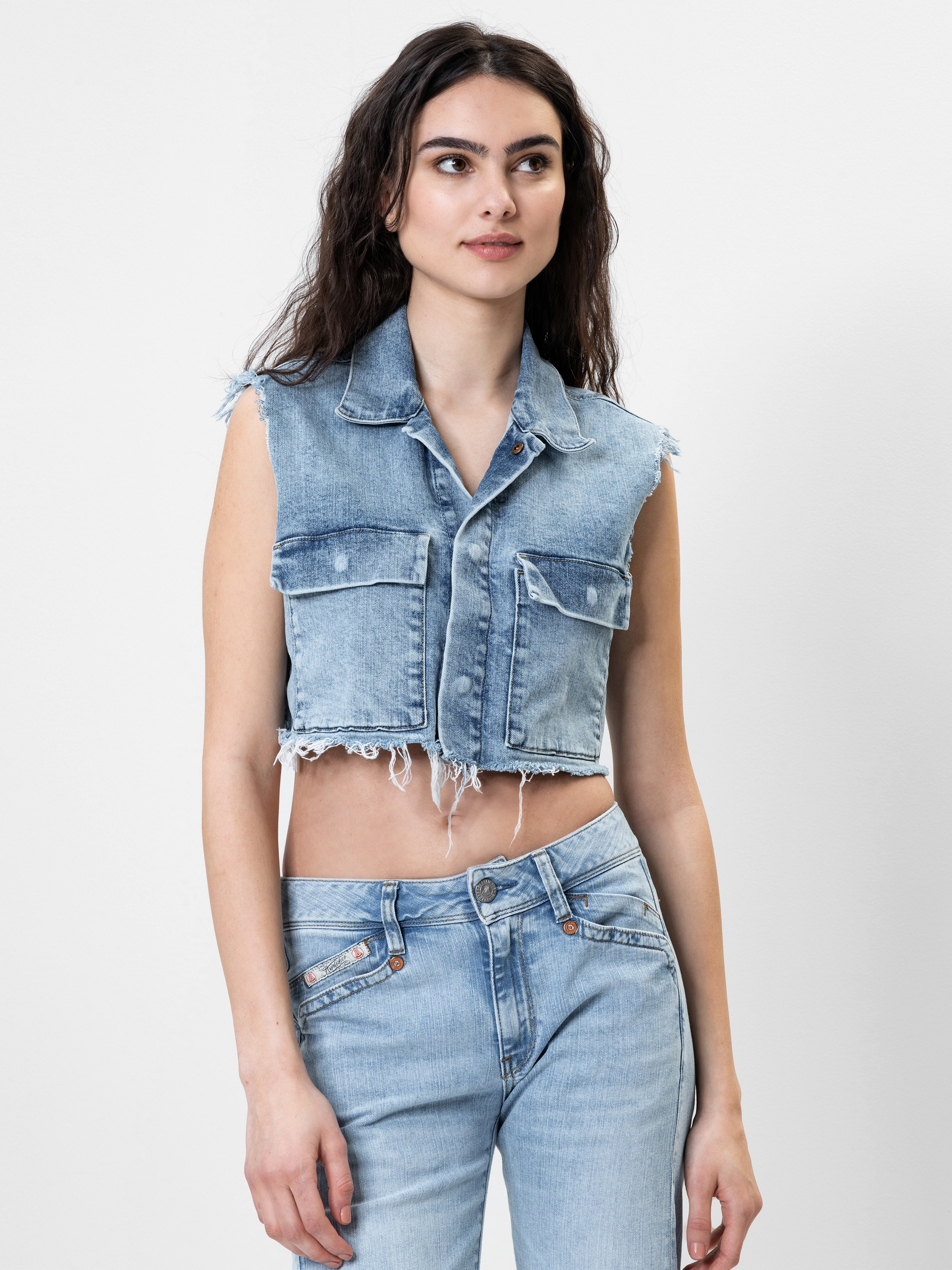 Eine Frau mit langen, gewellten dunklen Haaren trägt eine ärmellose, ausgefranste Jeansweste und hellblaue Jeans vor einem schlichten weißen Hintergrund.
