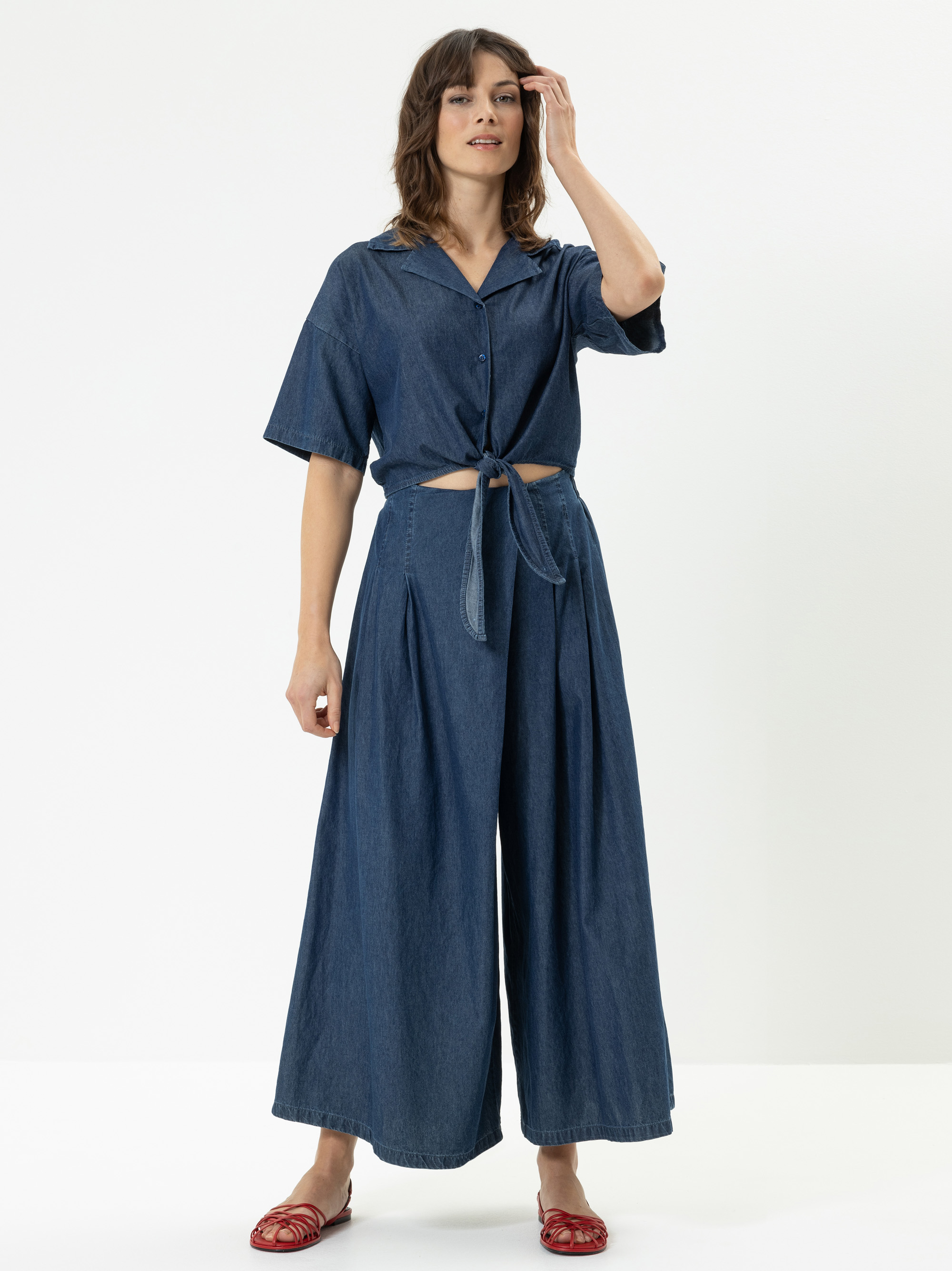 Eine Frau steht in einem Atelier und trägt ein blaues, kurzärmeliges, in der Taille geknöpftes Jeansoberteil und eine dazu passende, weit geschnittene Jeanshose. Sie hat schulterlanges braunes Haar und trägt rote Sandalen.