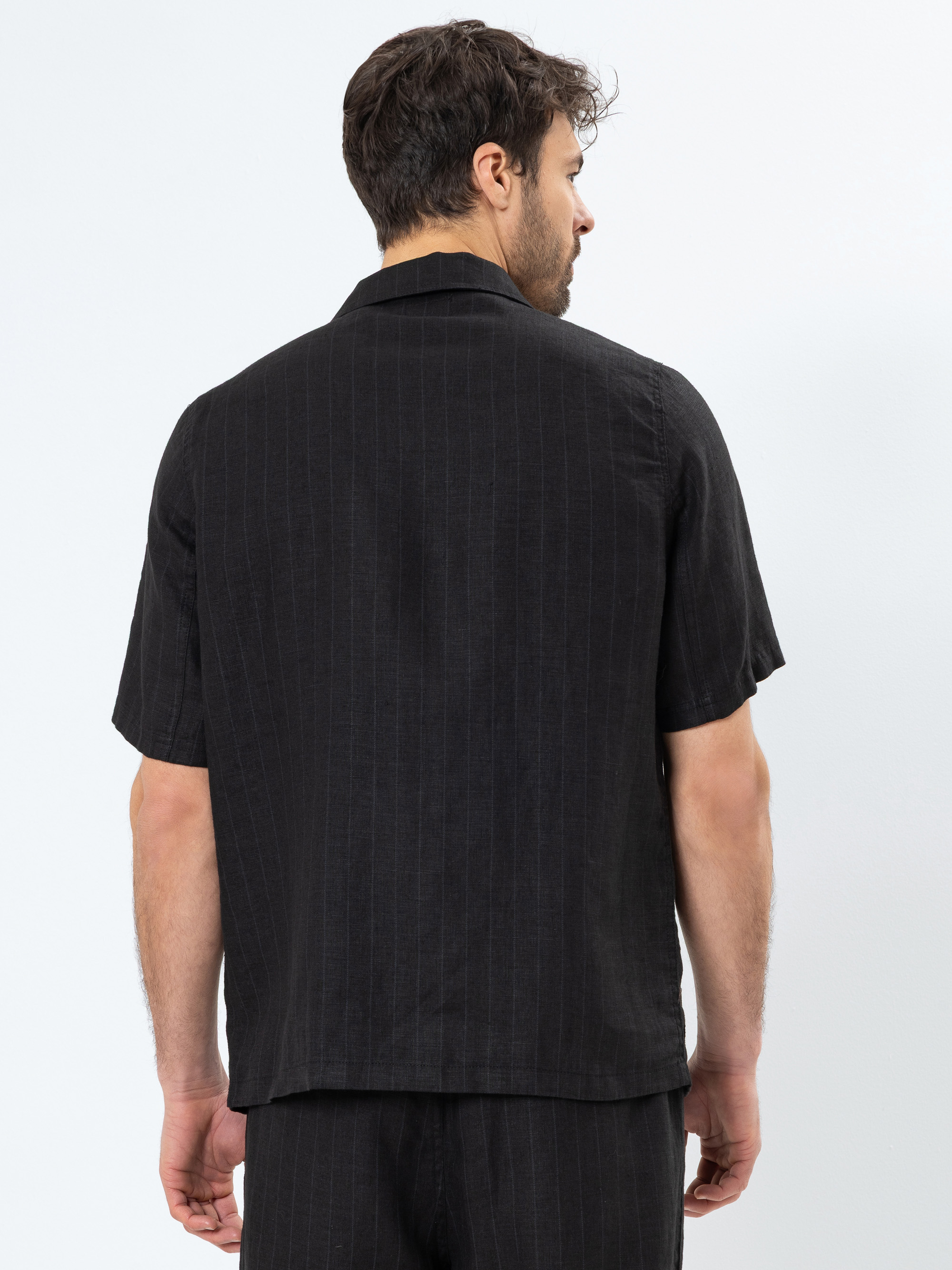 Ein Mann mit kurzen braunen Haaren steht mit dem Rücken zur Kamera und trägt ein schwarzes kurzärmeliges Button-up-Hemd und eine passende schwarze Hose. Das Hemd hat ein dezentes vertikales Nadelstreifenmuster.