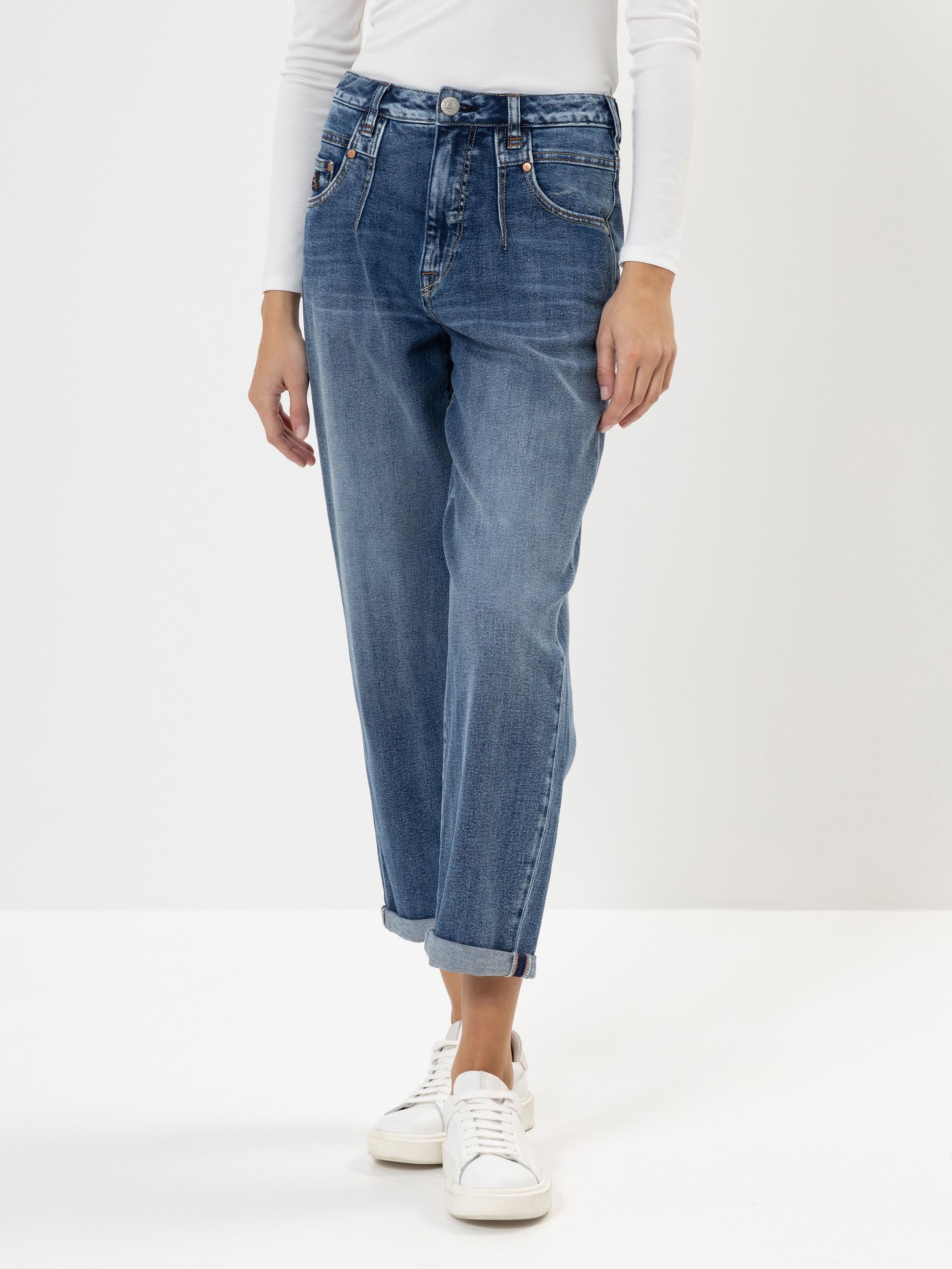 Eine Person, die ein weißes, langärmeliges Oberteil, blaue, hochgekrempelte Jeans und weiße Turnschuhe trägt, steht vor einem schlichten weißen Hintergrund. Der Kopf der Person ist auf dem Bild nicht zu sehen.