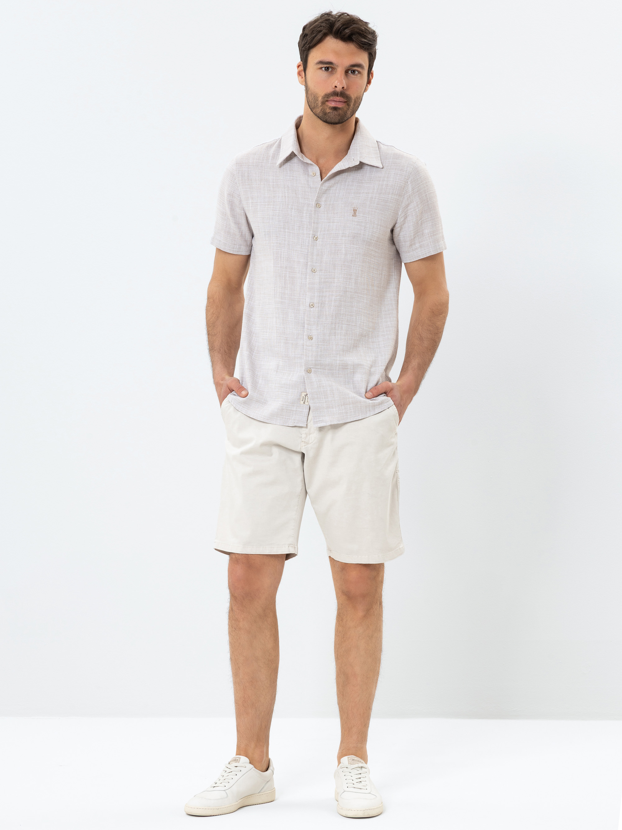 Ein Mann steht vor einem weißen Hintergrund, trägt ein hellgraues, kurzärmeliges Button-up-Hemd, beigefarbene Shorts und weiße Turnschuhe und hat die Hände in den Taschen.