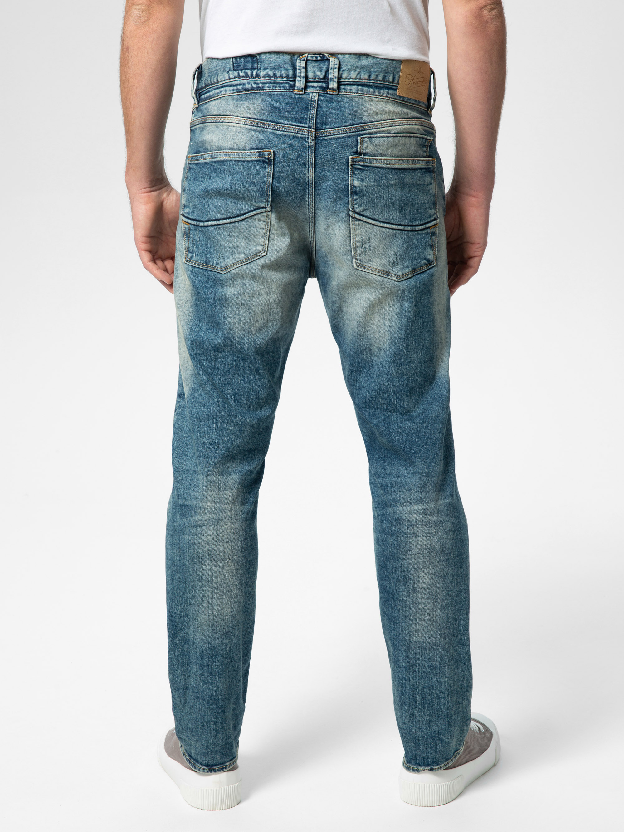 Eine Person steht mit dem Gesicht nach außen, trägt eine verblichene blaue Jeans, ein weißes T-Shirt und weiße Turnschuhe vor einem schlichten, hellen Hintergrund.