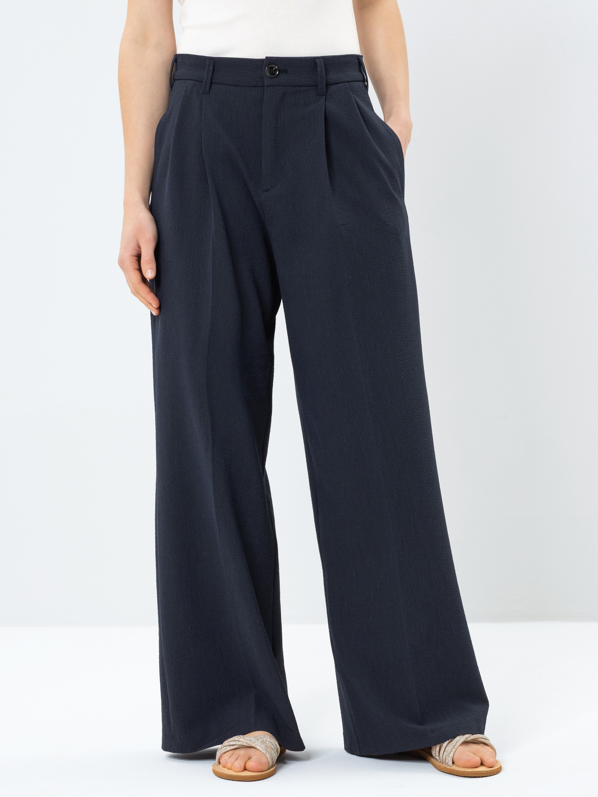 Eine Person, die eine marineblaue Hose mit hoher Taille und weitem Bein sowie Sandalen trägt und die Hände in den Taschen hat, steht vor einem schlichten hellen Hintergrund.