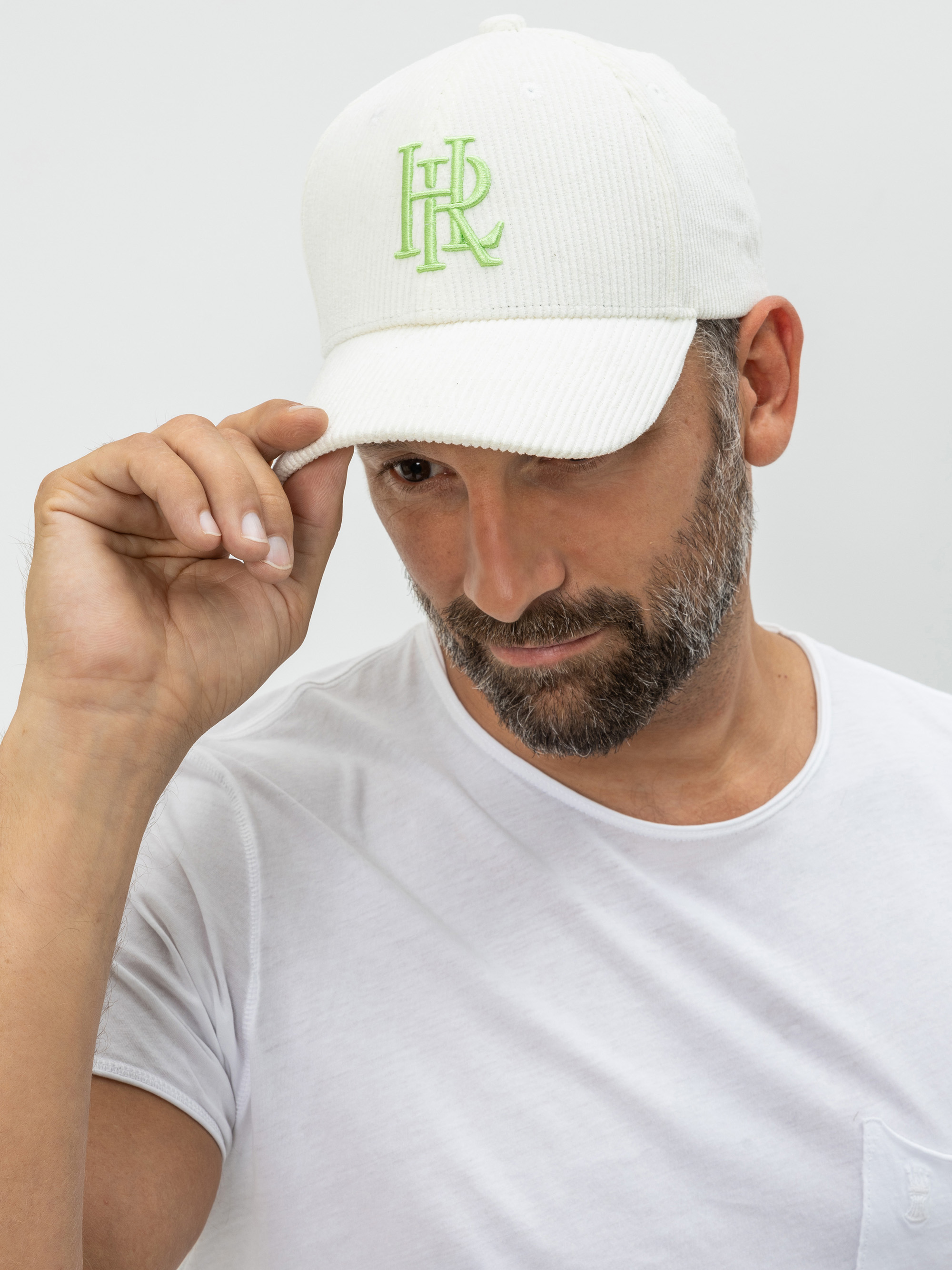 Ein Mann mit Bart und grauen Haaren, der ein weißes T-Shirt und eine weiße Baseballmütze mit grünem HR-Logo trägt, blickt nach unten und hält mit einer Hand den Rand seiner Mütze.