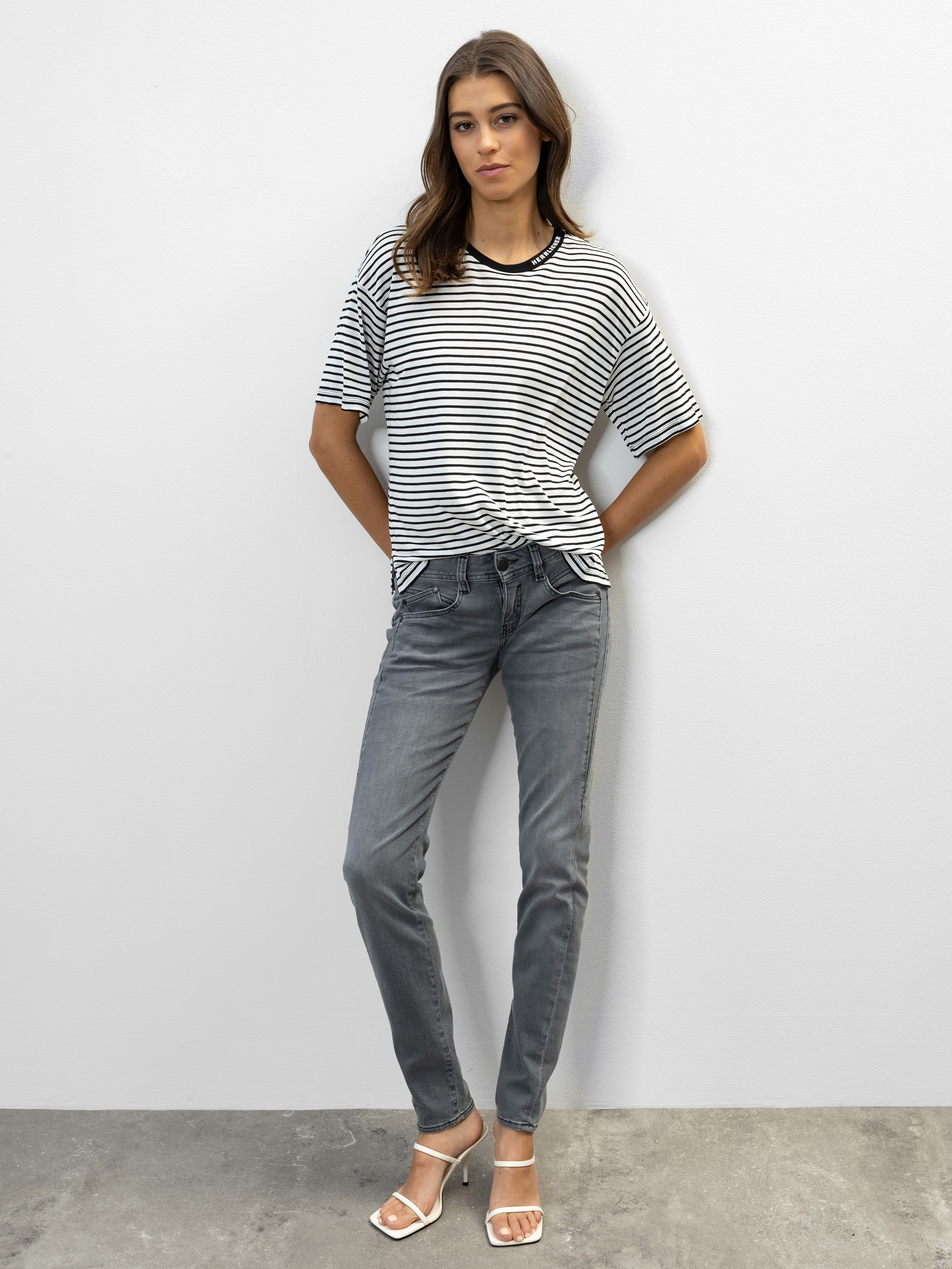 Eine Frau steht vor einer schlichten weißen Wand, trägt ein schwarz-weiß gestreiftes T-Shirt, graue Jeans und weiße Sandalen mit Absatz. Sie hat langes, offenes Haar und ihre Hände stecken in den Taschen.