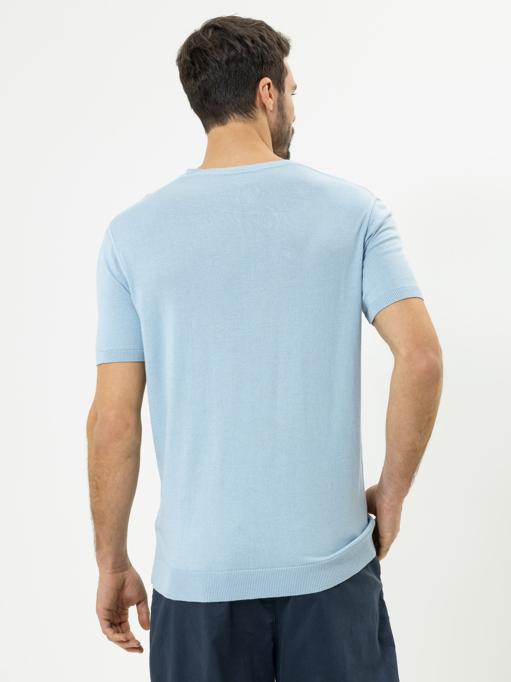 Ein Mann steht mit dem Rücken zur Kamera, trägt einen hellblauen, kurzärmeligen Pullover und dunkle Shorts vor einem schlichten weißen Hintergrund.