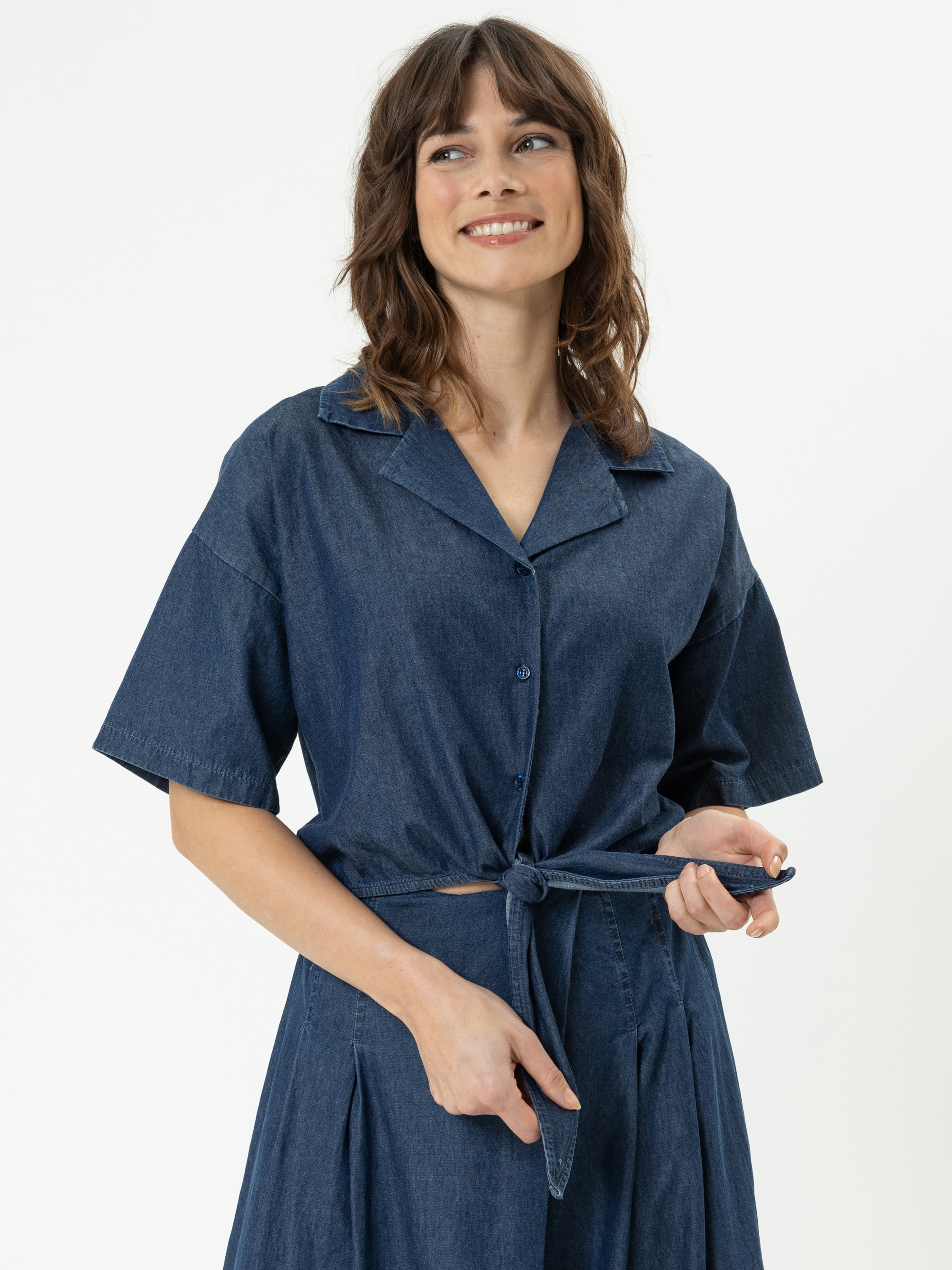 Eine Frau mit gewelltem braunem Haar lächelt, während sie ein kurzärmeliges, hochgeknöpftes Jeanskleid trägt und einen Gürtel um ihre Taille bindet. Sie steht vor einem schlichten weißen Hintergrund.