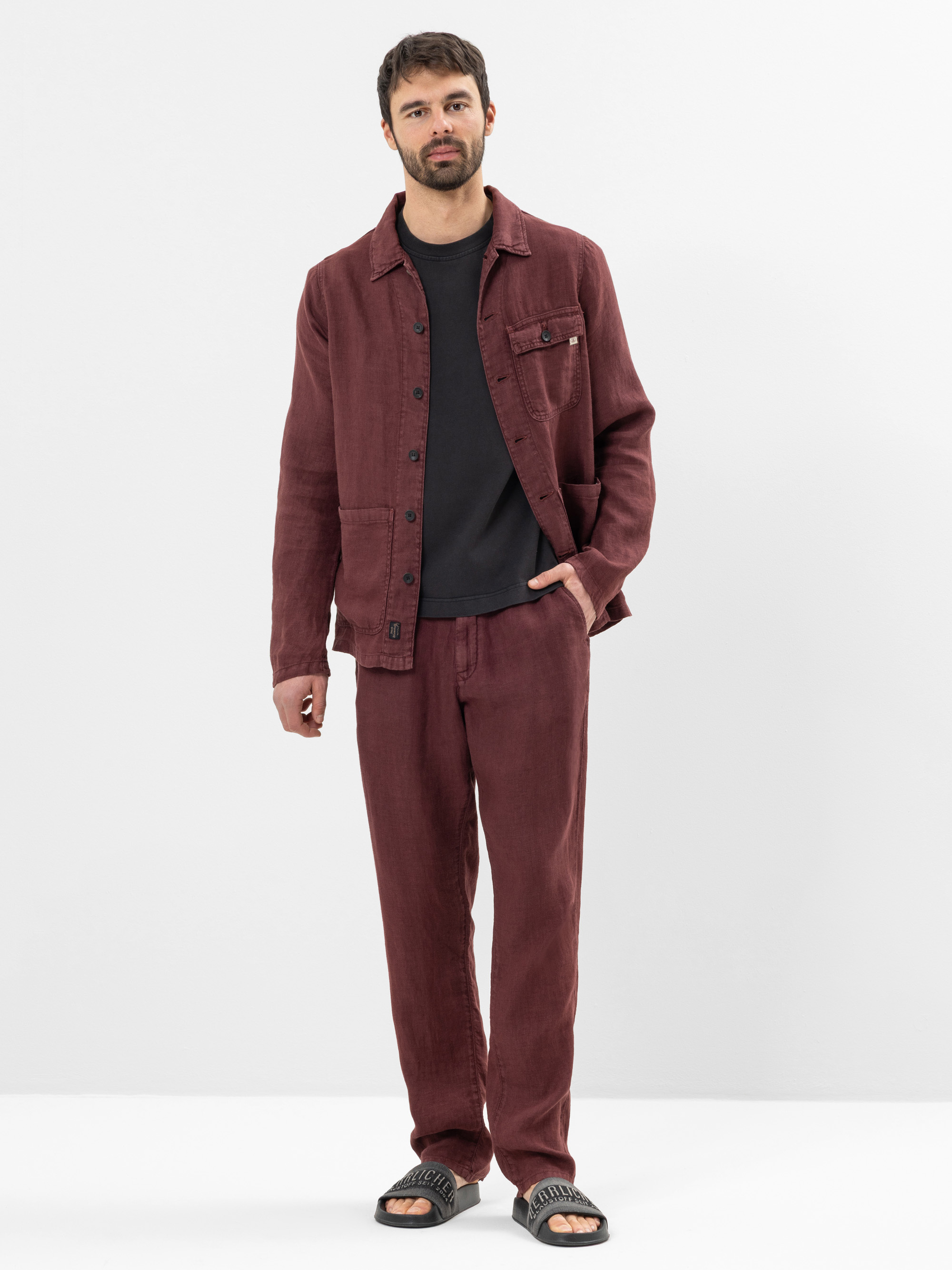 Ein Mann steht vor einem schlichten weißen Hintergrund und trägt eine kastanienbraune Button-up-Jacke, eine passende kastanienbraune Hose, ein schwarzes T-Shirt und schwarze Slide-Sandalen. Er hat kurze Haare, einen Bart und einen entspannten Gesichtsau