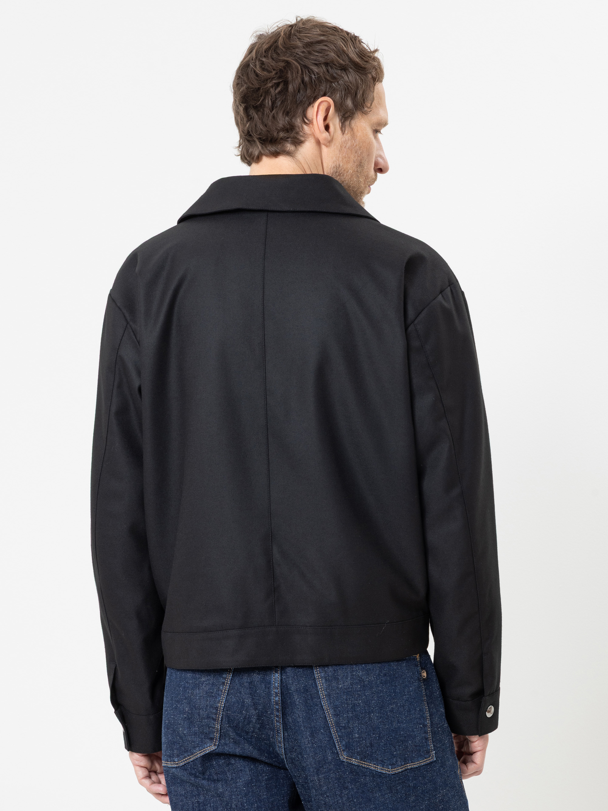 Vor einem schlichten weißen Hintergrund steht ein Mann mit kurzen braunen Haaren, der mit dem Rücken zur Kamera steht und eine schwarze Jacke sowie blaue Jeans trägt.