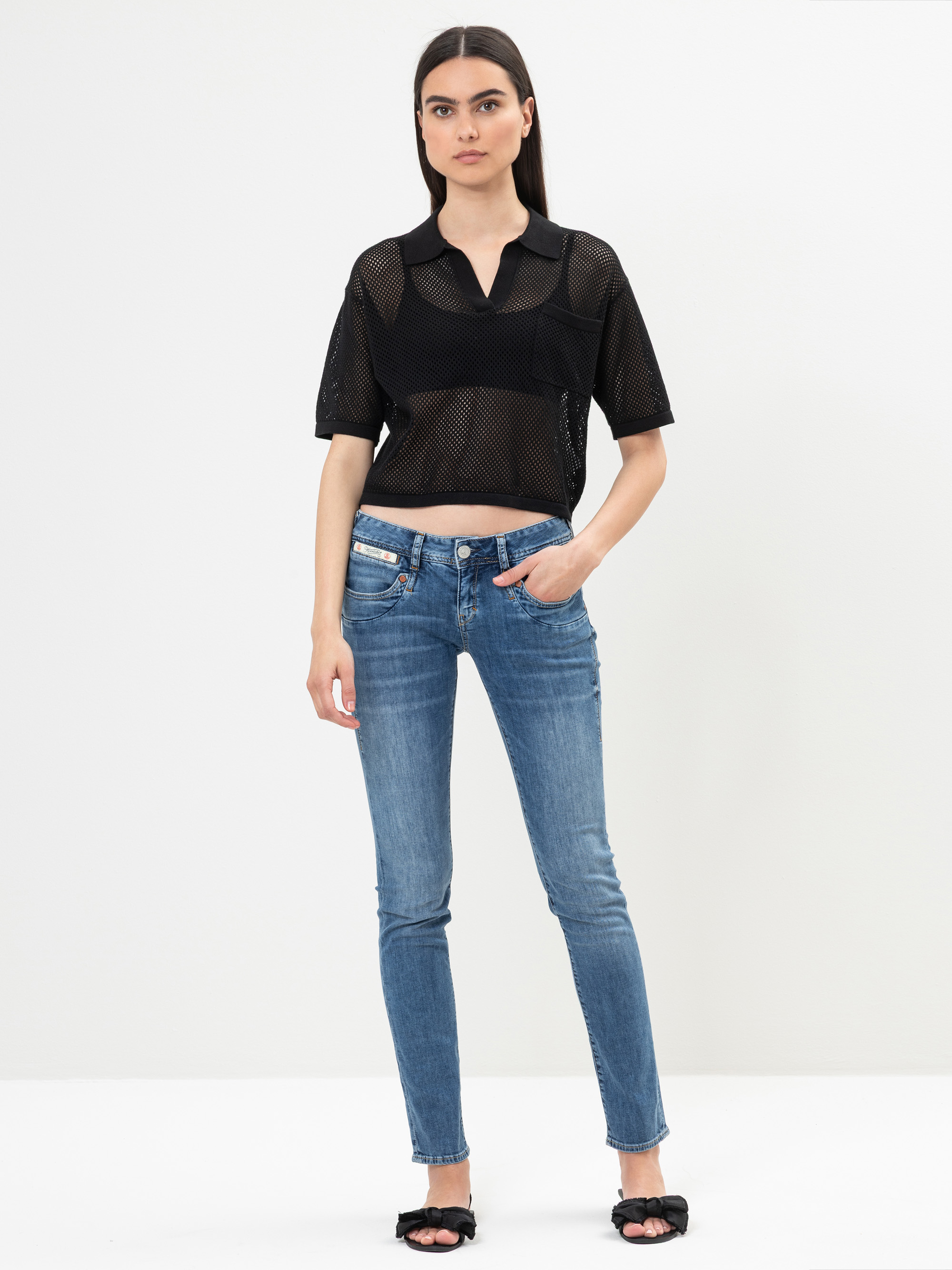 Eine Person steht vor einem schlichten weißen Hintergrund und trägt ein schwarzes, kurzärmeliges Mesh-Crop-Top, blaue Skinny-Jeans mit Reißverschluss und schwarze, offene Absätze. Eine Hand steckt in der Hosentasche und sie hat langes dunkles Haar.